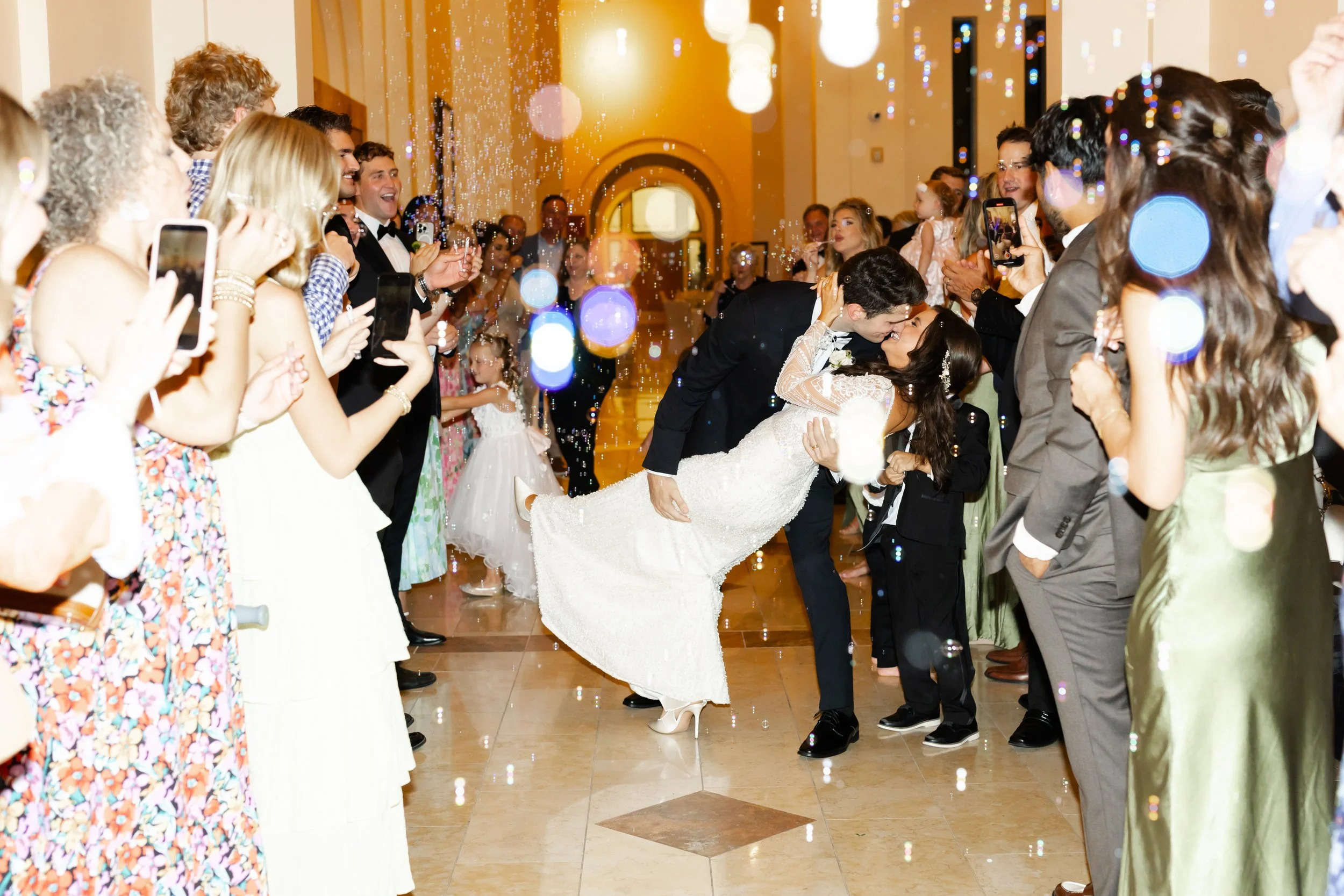 A newlywed couple kissing while being surrounded by guests at their wedding reception. The bride is leaning back in the groom's arms as they share a kiss, with guests cheering and taking photos by Wild Ivy Creative photography