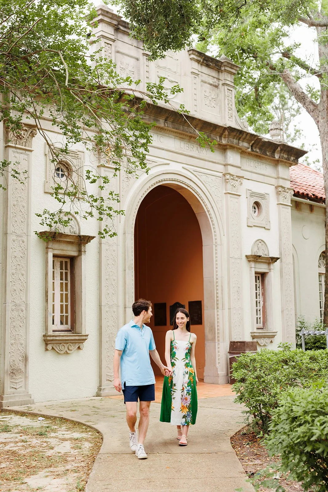 Couple walking together in front of a historic Houston library surrounded by greenery during a sunny engagement session with Wild Ivy Creative.