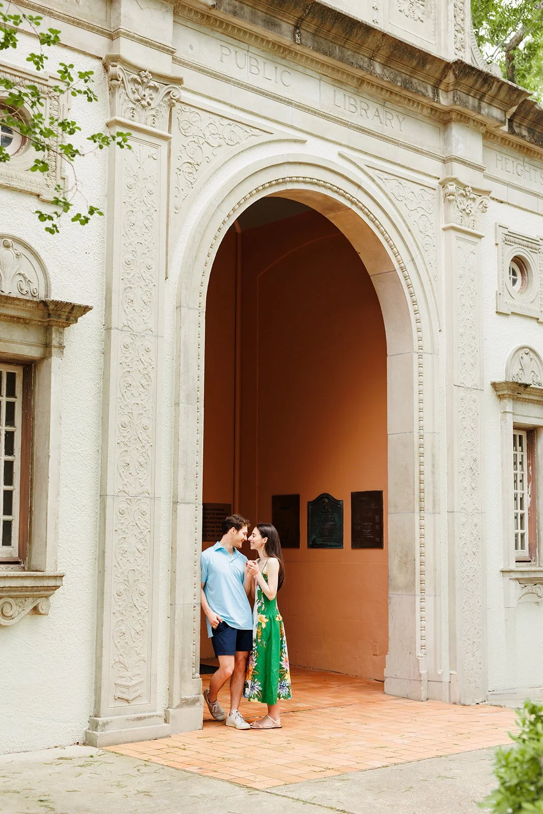A young couple standing close together and holding hands outside a historic library building, smiling at each other.