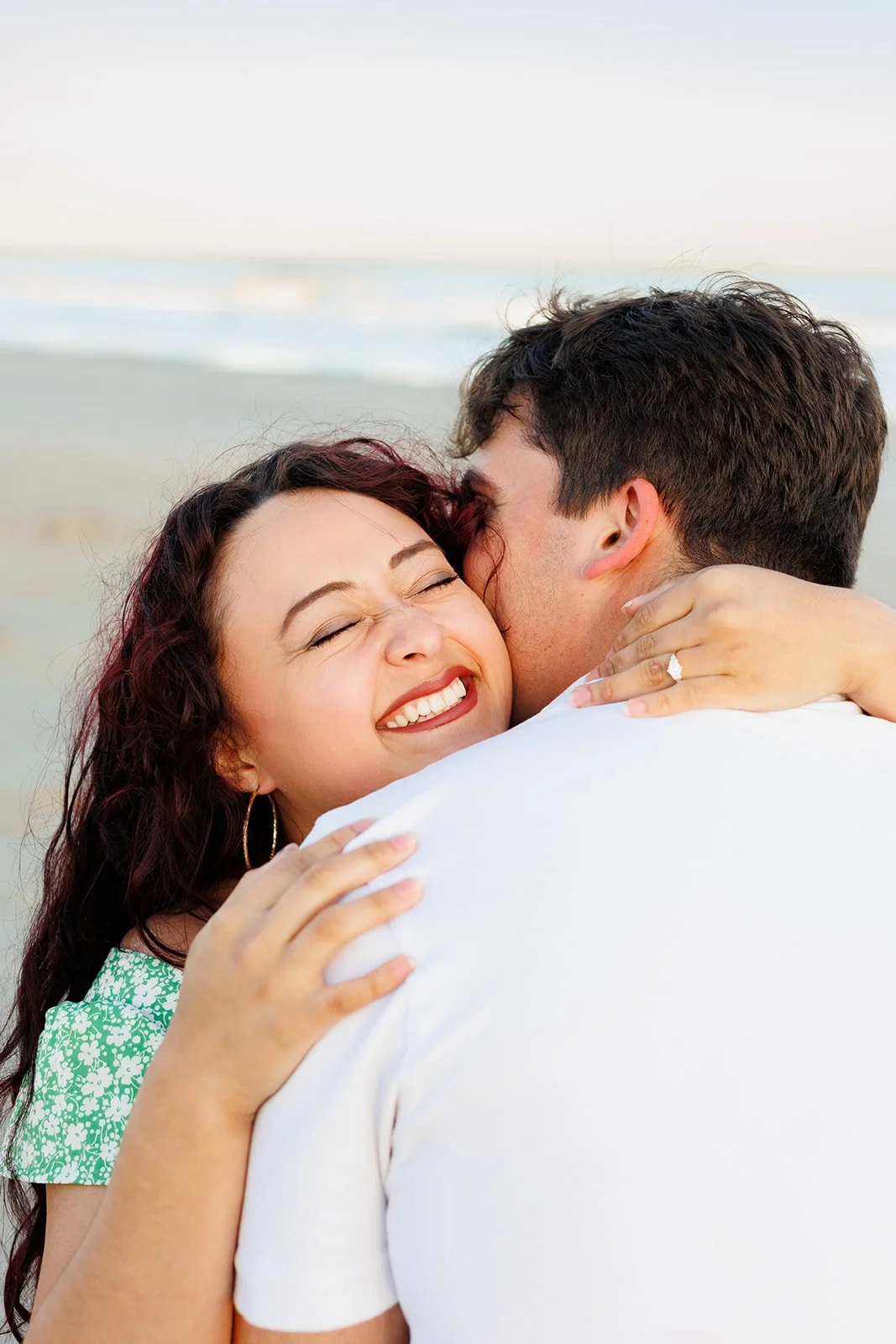 A happy woman with curly red hair and hoop earrings hugging a man with dark hair on the beach, both smiling and showing affection.