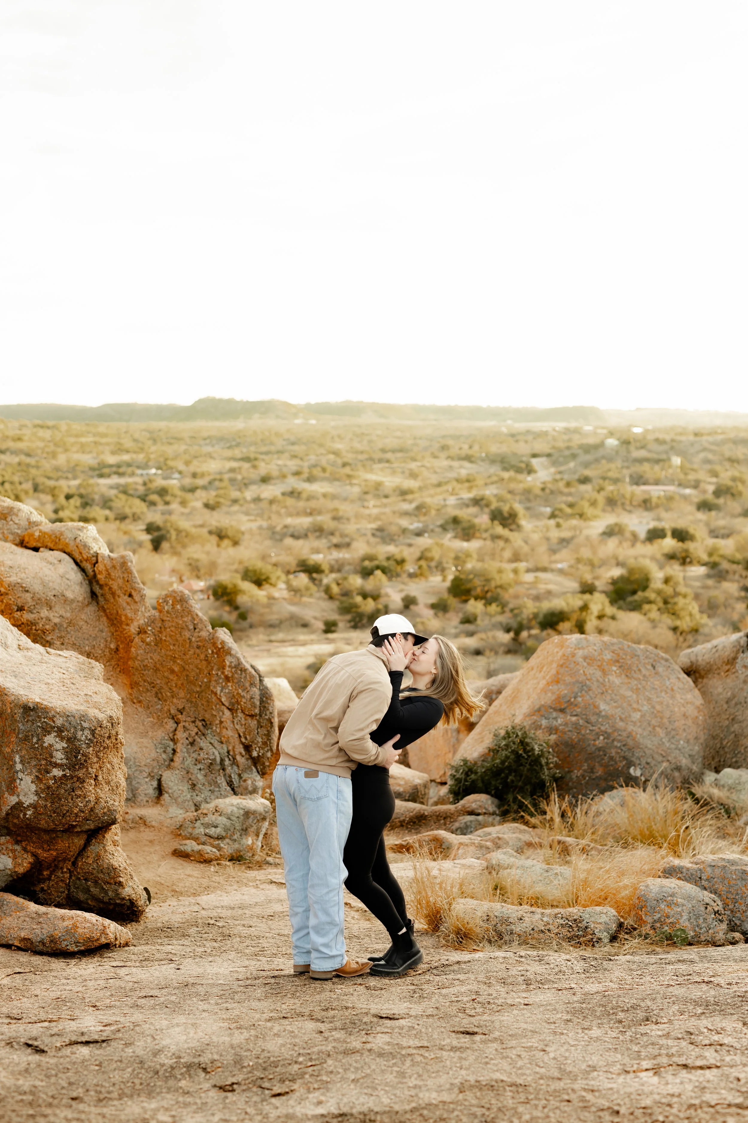 A couple sharing a kiss on a dirt path surrounded by large rocks in a desert landscape at sunset.