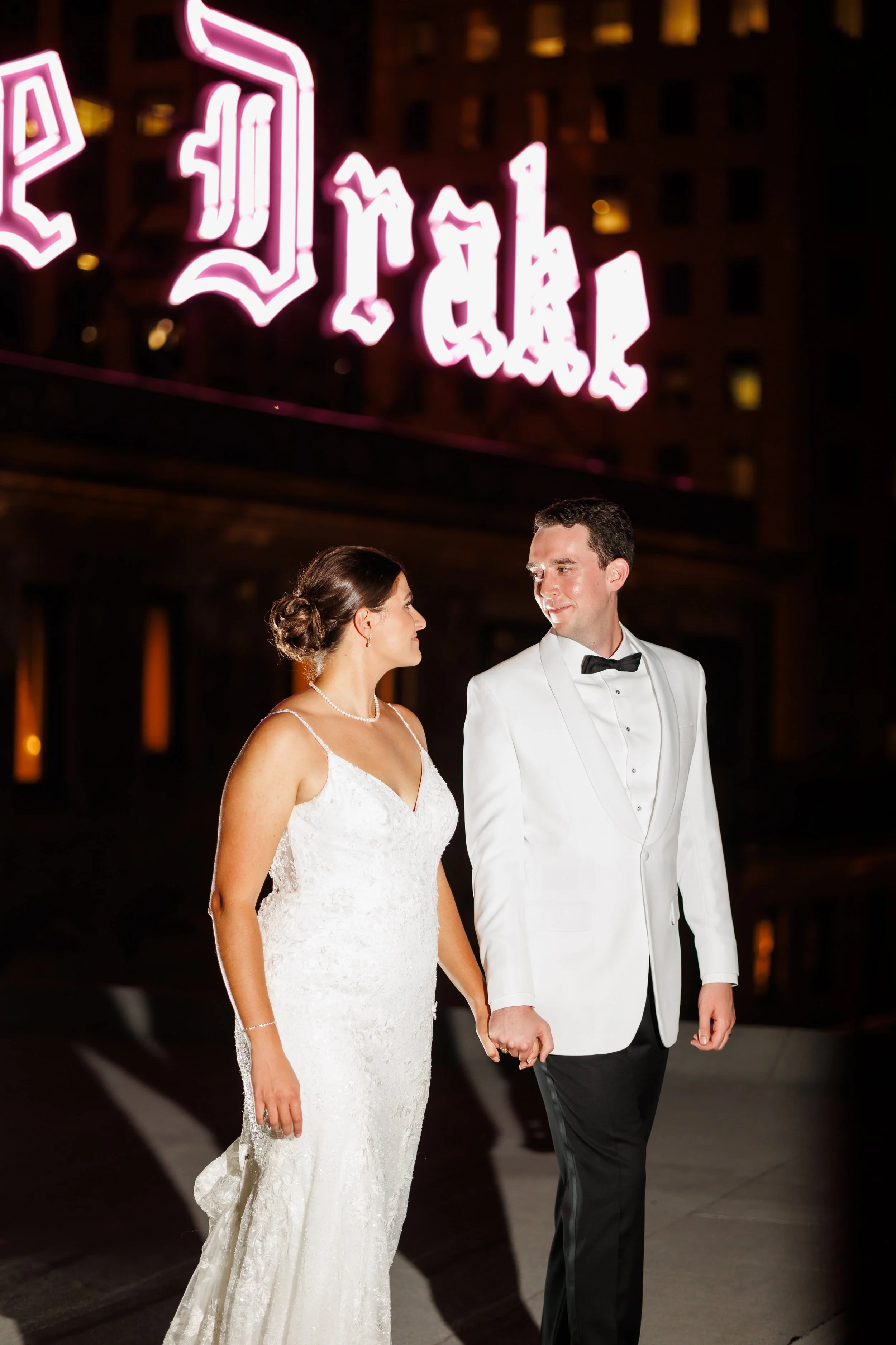Newlywed couple holding hands outside a Houston reception venue at night, dressed in wedding attire, with a neon pink “Drybar” sign glowing in the background.