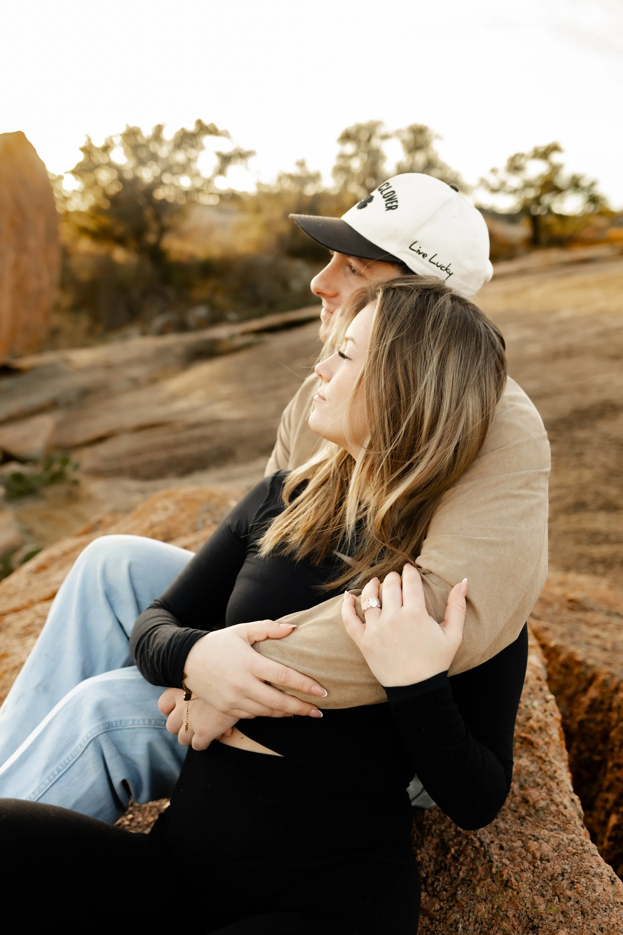 A young couple sitting outdoors on rocks, embracing each other with their eyes closed, during sunset in a natural landscape at Enchanted Rock for an engagement session
