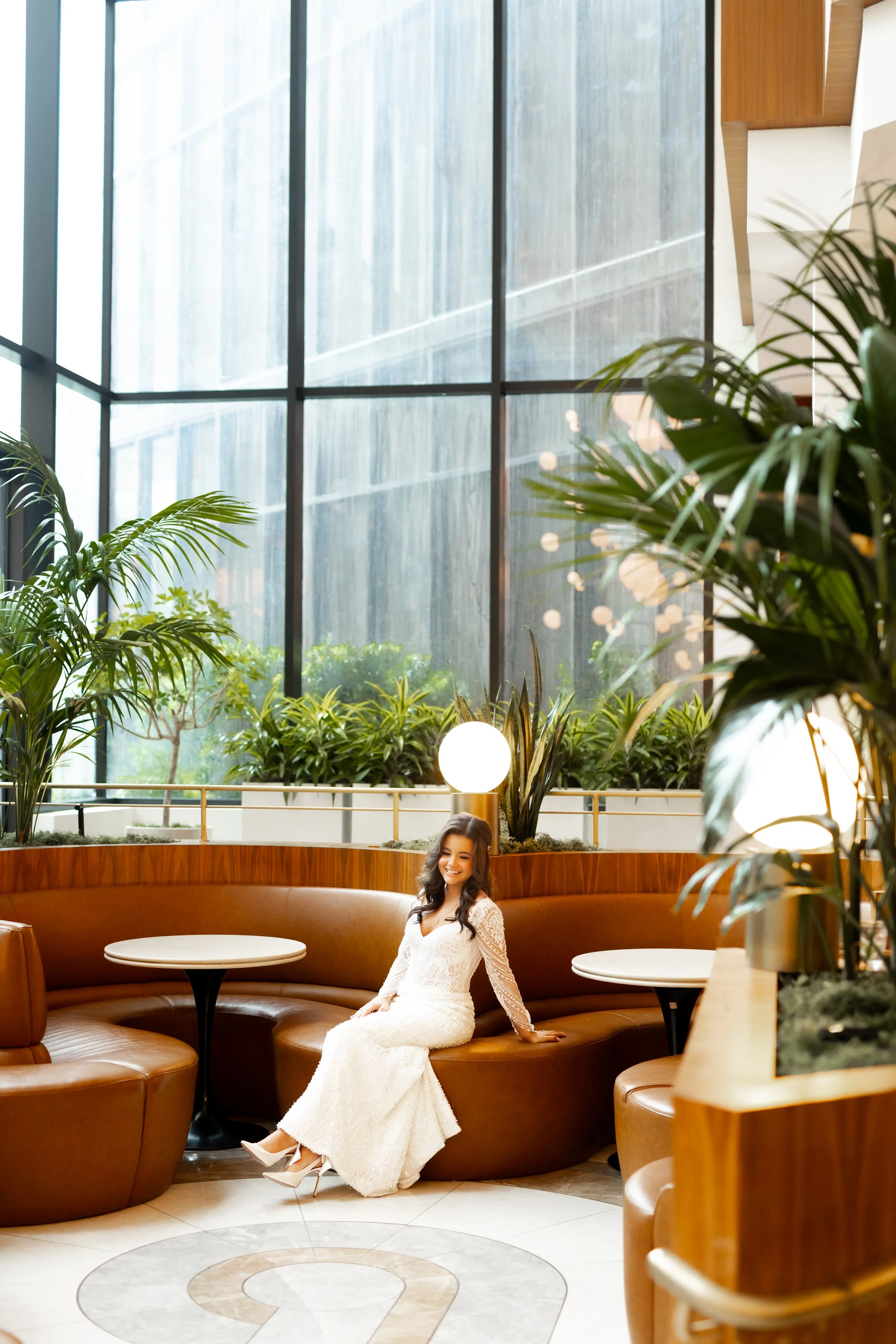 A woman in a white lace dress sitting on a curved leather sofa inside a modern building with large windows, plants, and decorative lighting captured by top rated Houston wedding photographer. 