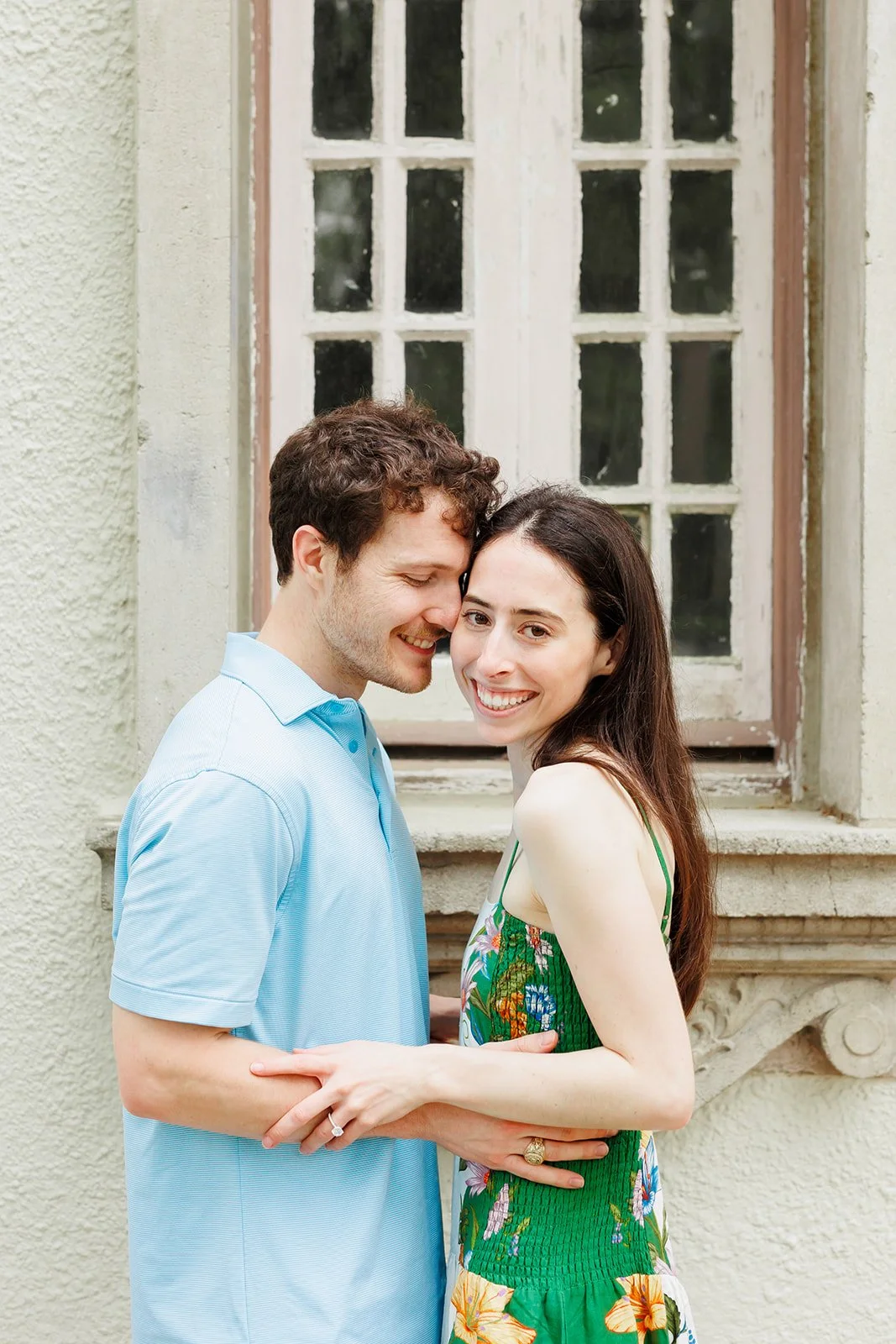 A happy couple embraces in front of an old window, smiling and touching foreheads, with the man wearing a light blue shirt and the woman in a colorful green dress during a Houston Heights Surprise Proposal.
