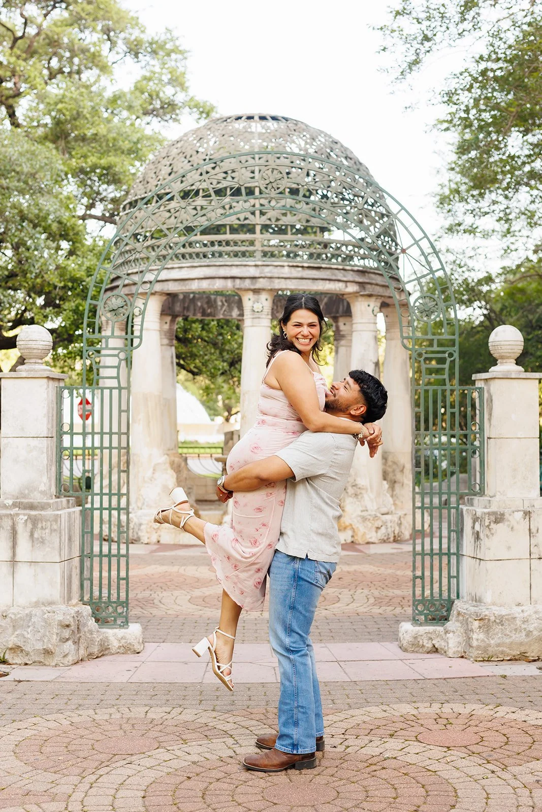 Joyful park proposal or engagement session with a man lifting his partner in a pink floral dress near a gazebo in Hermann Park in Houston — captured by Wild Ivy Creative.