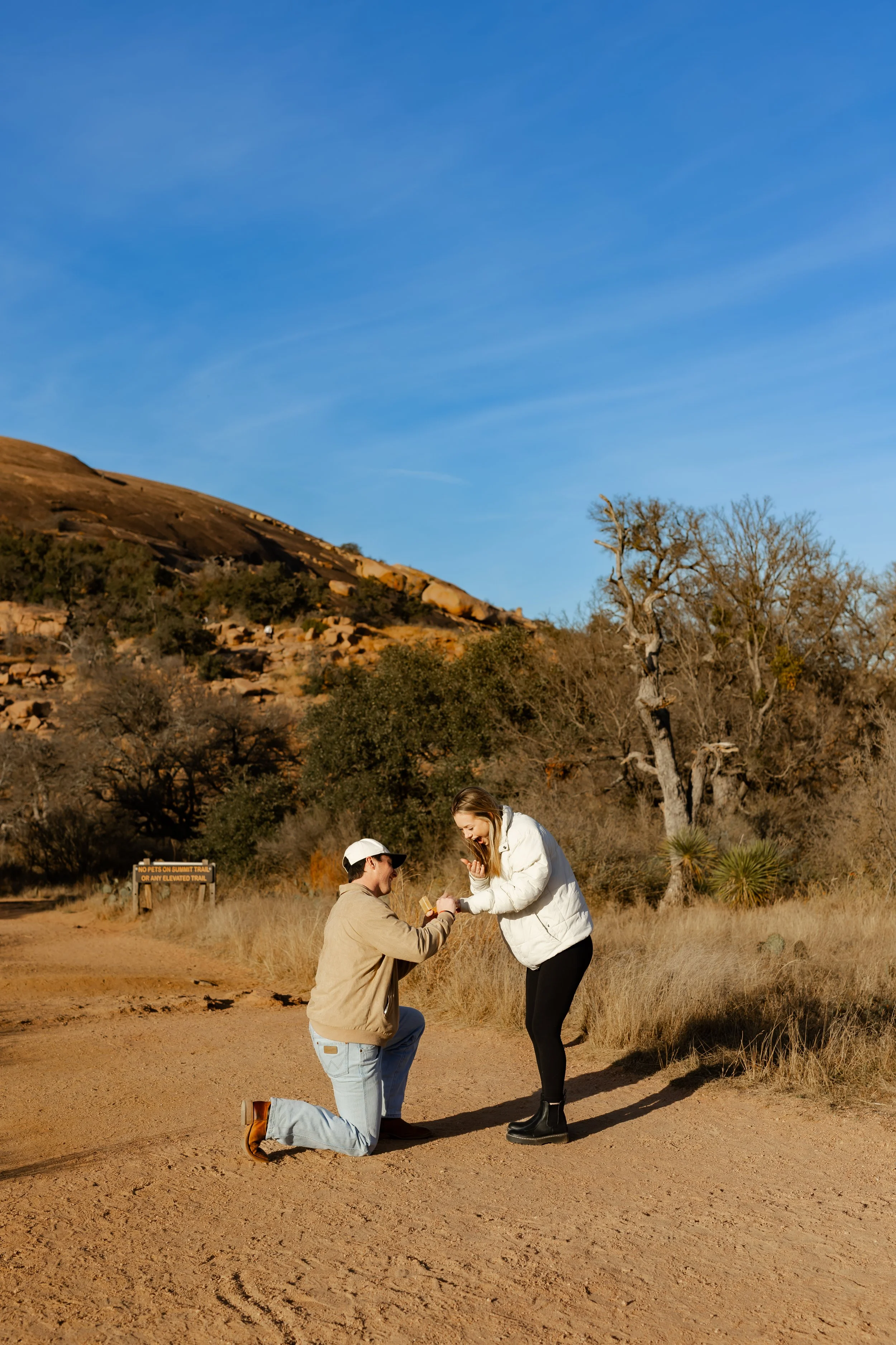 A man proposing marriage, at Enchanted Rock in Texas, to a woman in a desert landscape with trees and hills in the background during sunset.