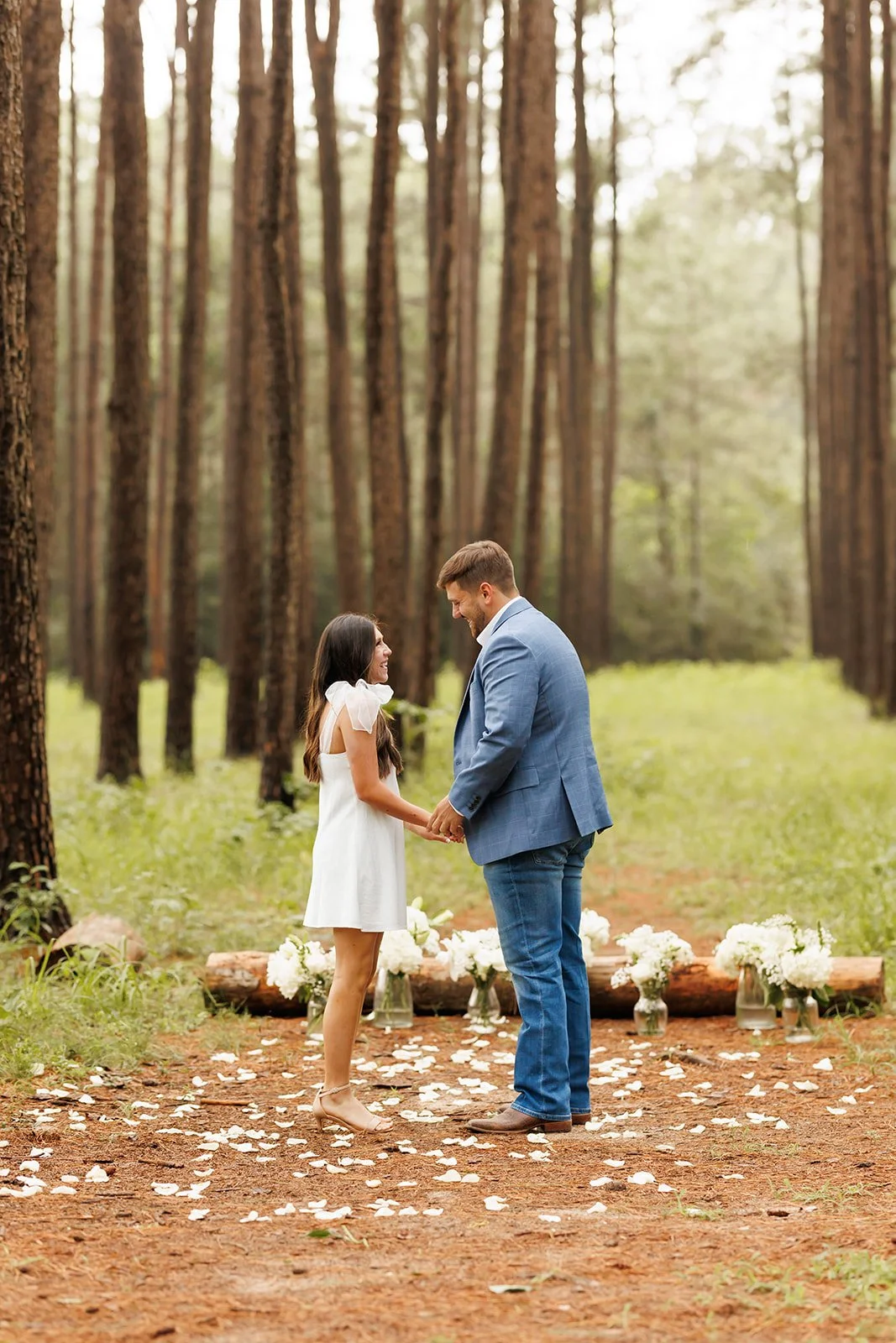 A couple holding hands and smiling during their outdoor wedding ceremony in a forest, with flower arrangements and white rose petals on the ground.