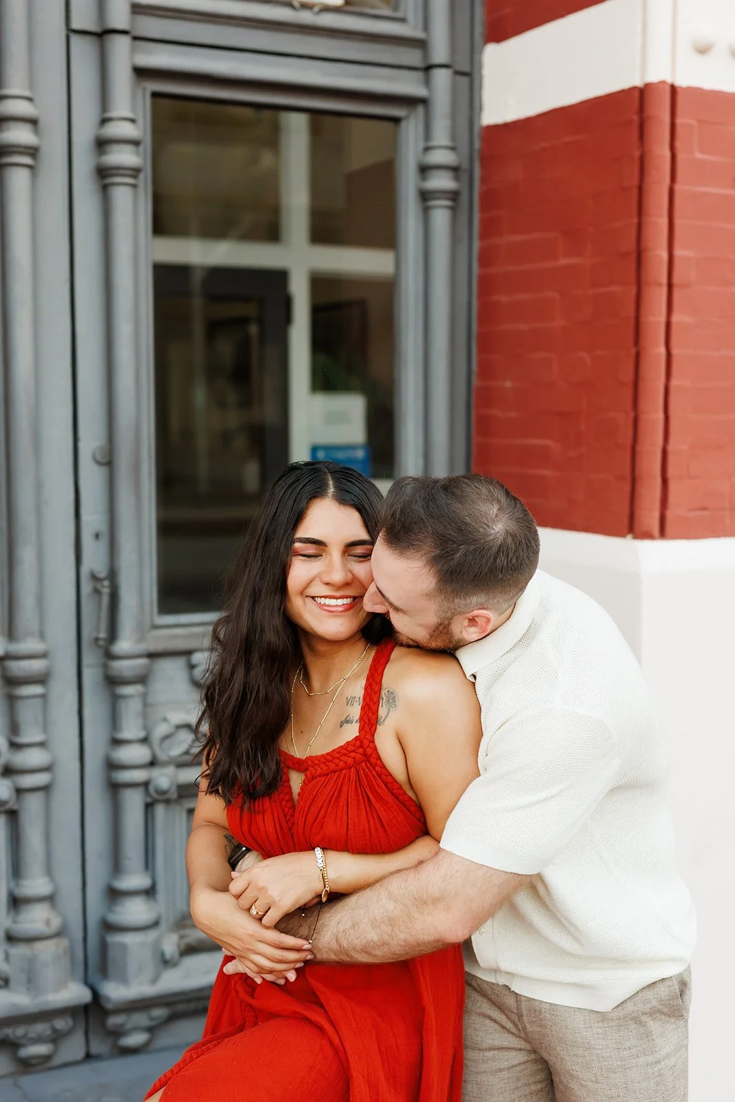 Intimate and joyful Houston engagement moment — woman in red dress embracing her partner near a gray door and red brick wall — captured by Wild Ivy Creative.