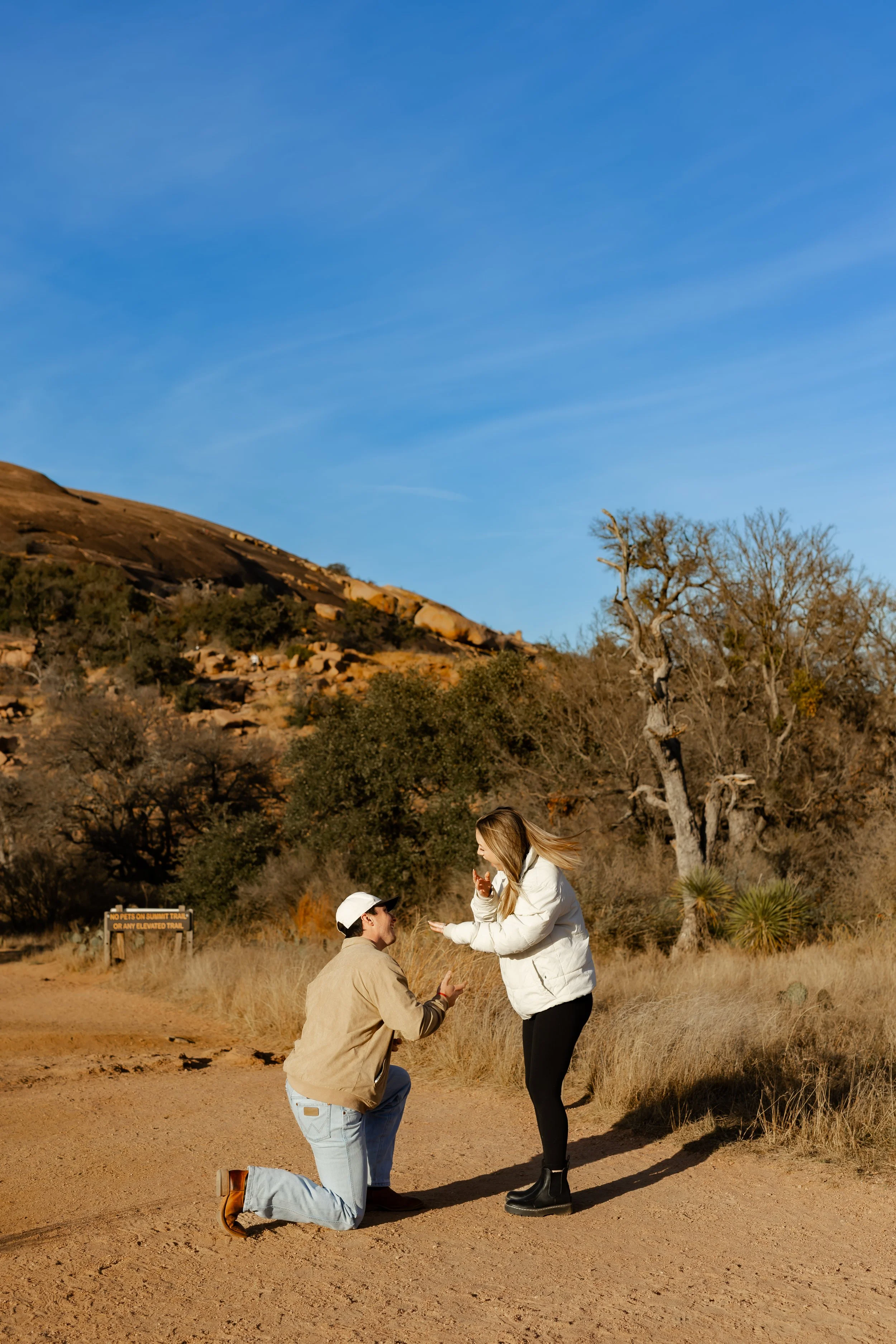 A man is kneeling with a woman standing in front of him on a dirt trail in a desert landscape, and they appear to be proposing or celebrating a special moment at Enchanted Rock State Park for a surprise proposal.