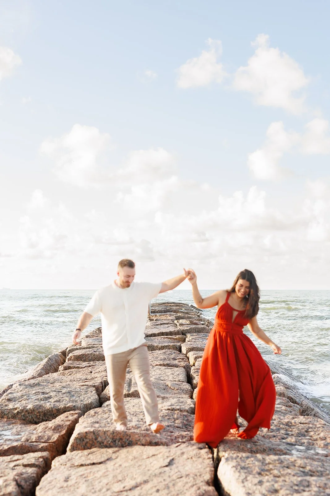 Dreamy Galveston beach engagement session on a rocky pier — couple walking hand-in-hand under a cloudy sky — Wild Ivy Creative.