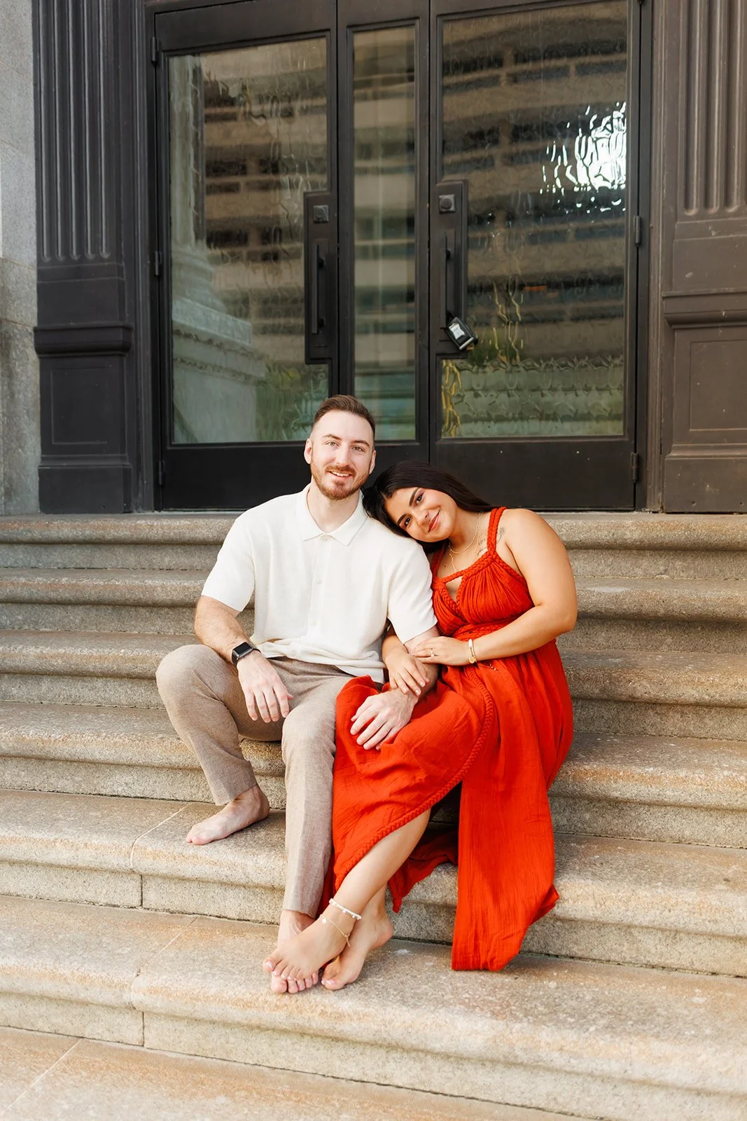 Sweet Houston engagement moment with a woman in red leaning on her partner’s shoulder on historic building steps — captured by Wild Ivy Creative.