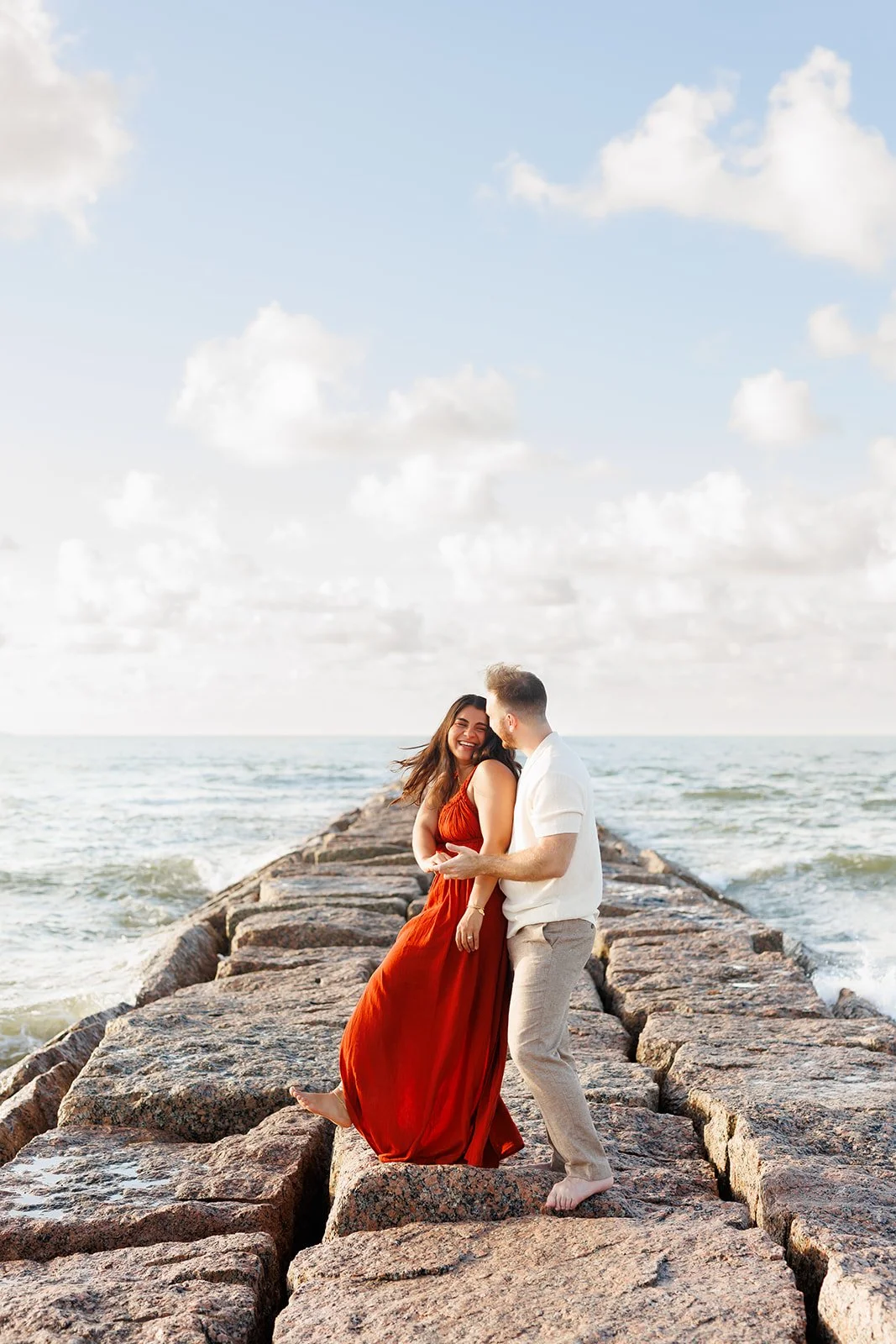 Vibrant Galveston engagement portrait — couple laughing on a rocky pier with waves and sky behind them — captured by Wild Ivy Creative.