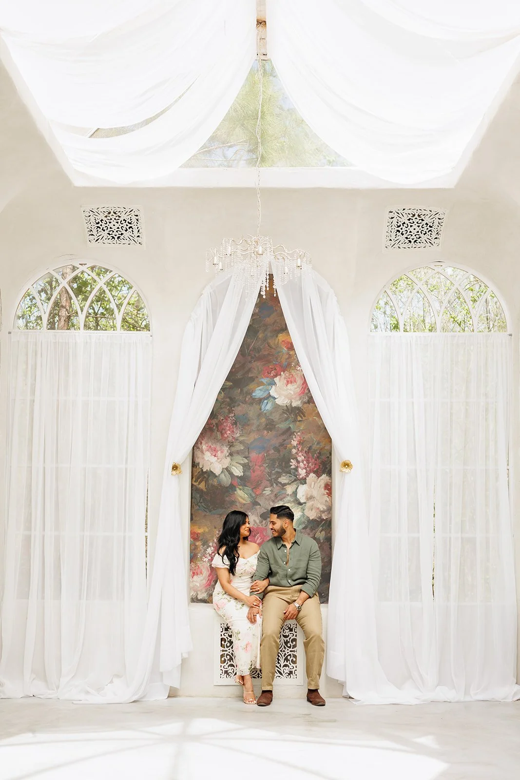 Indoor engagement session in a bright white room with floral decor, chandelier, and large windows, photographed by Wild Ivy Creative.