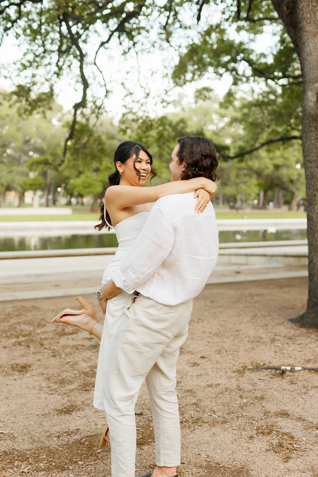 A man is lifting a woman in his arms in a park, both smiling at each other, surrounded by trees and a pond in the background in Houston during a Proposal and Engagement Session.