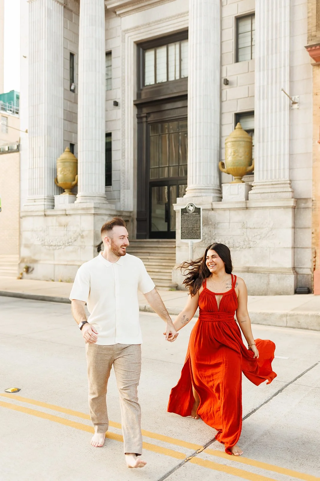 Vibrant Houston engagement photo with a barefoot couple walking in front of grand columns — woman in orange dress, man in beige — captured by Wild Ivy Creative.
