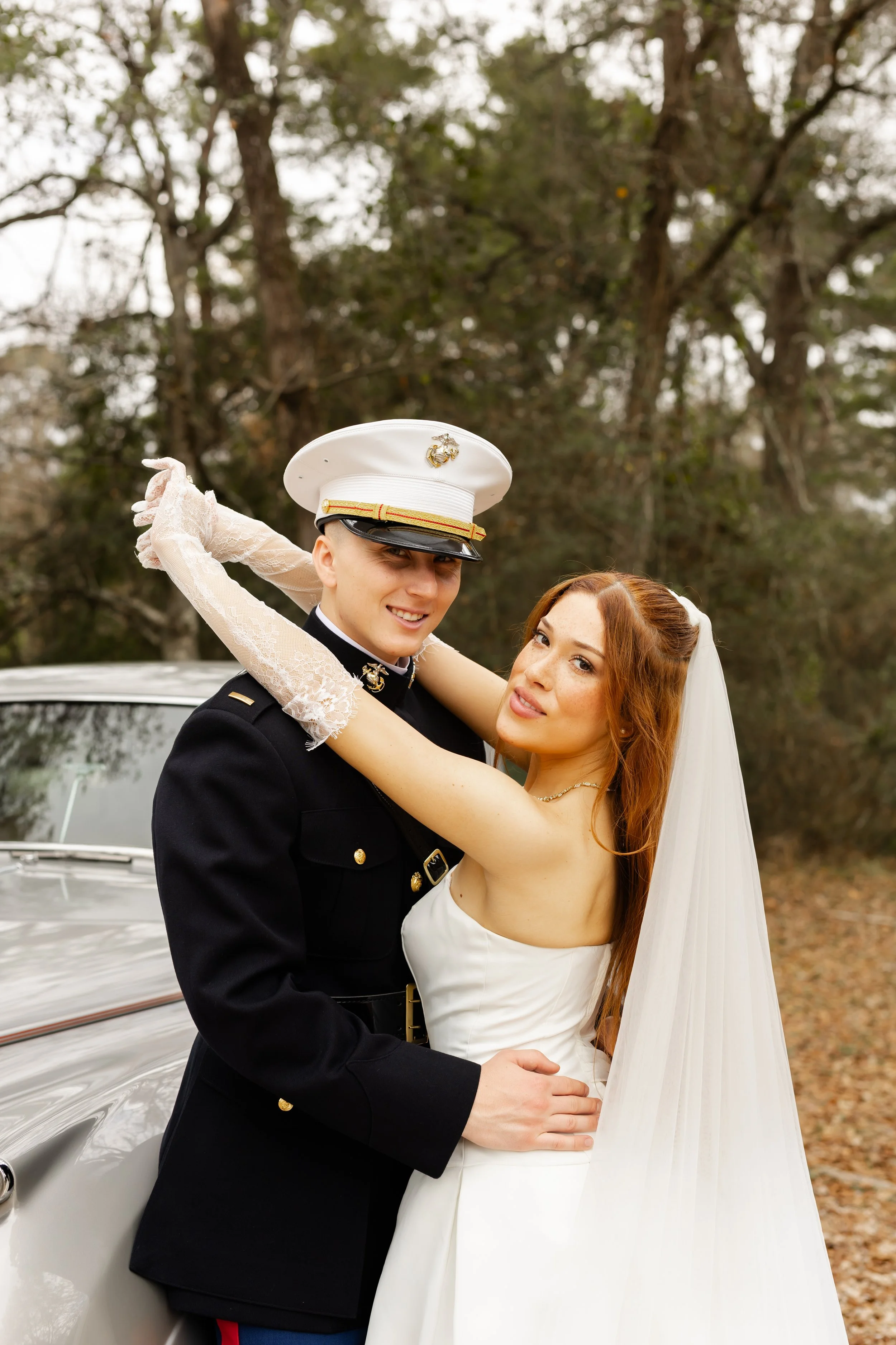 A newlywed couple, a woman with long red hair and a man in a military uniform, embracing outdoors with trees and a vintage car in the background at the Oak Atelier in the Woodlands photographed by Wild Ivy Creative Photography