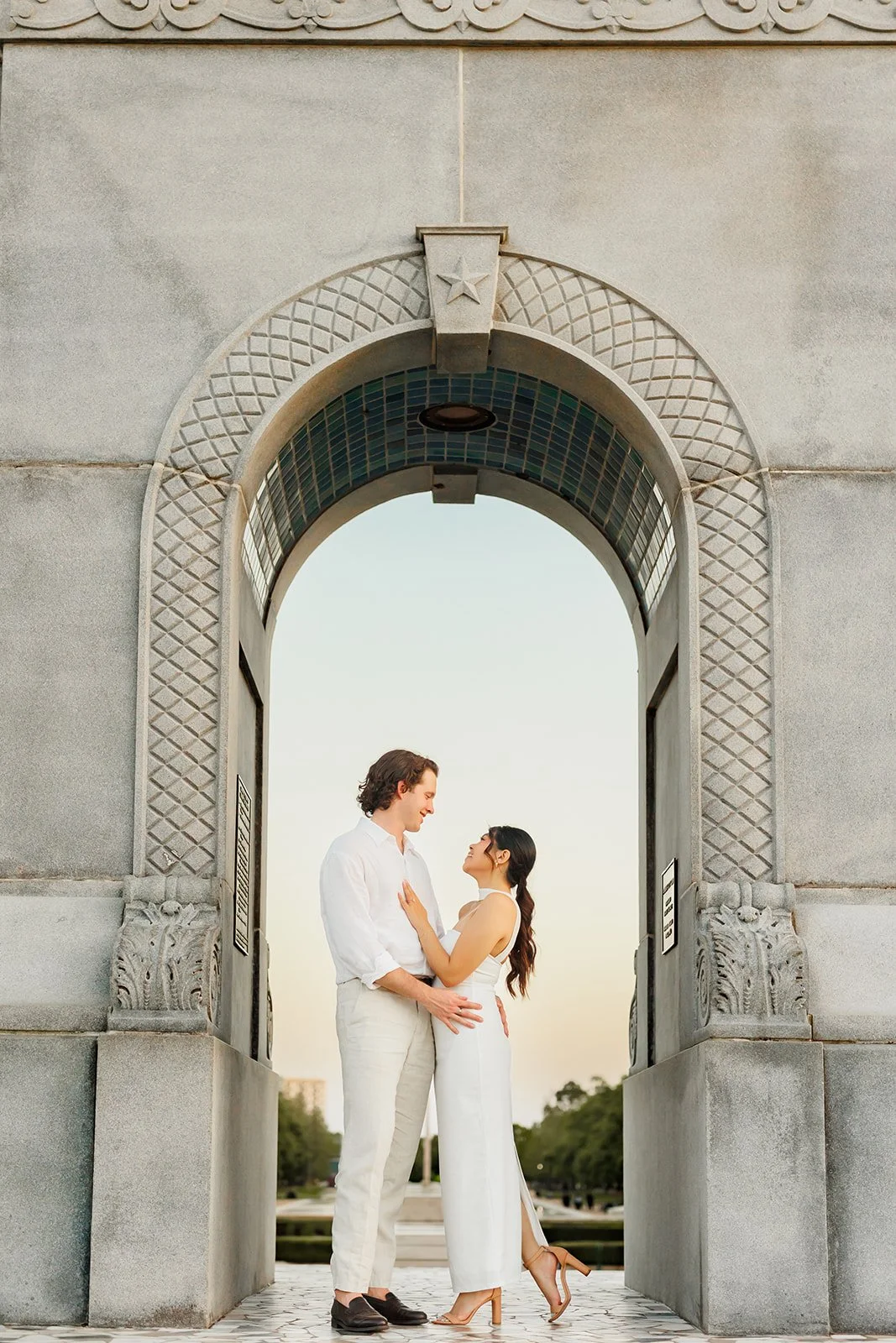 A couple standing in an archway, looking at each other affectionately, with a background of a park and sky.