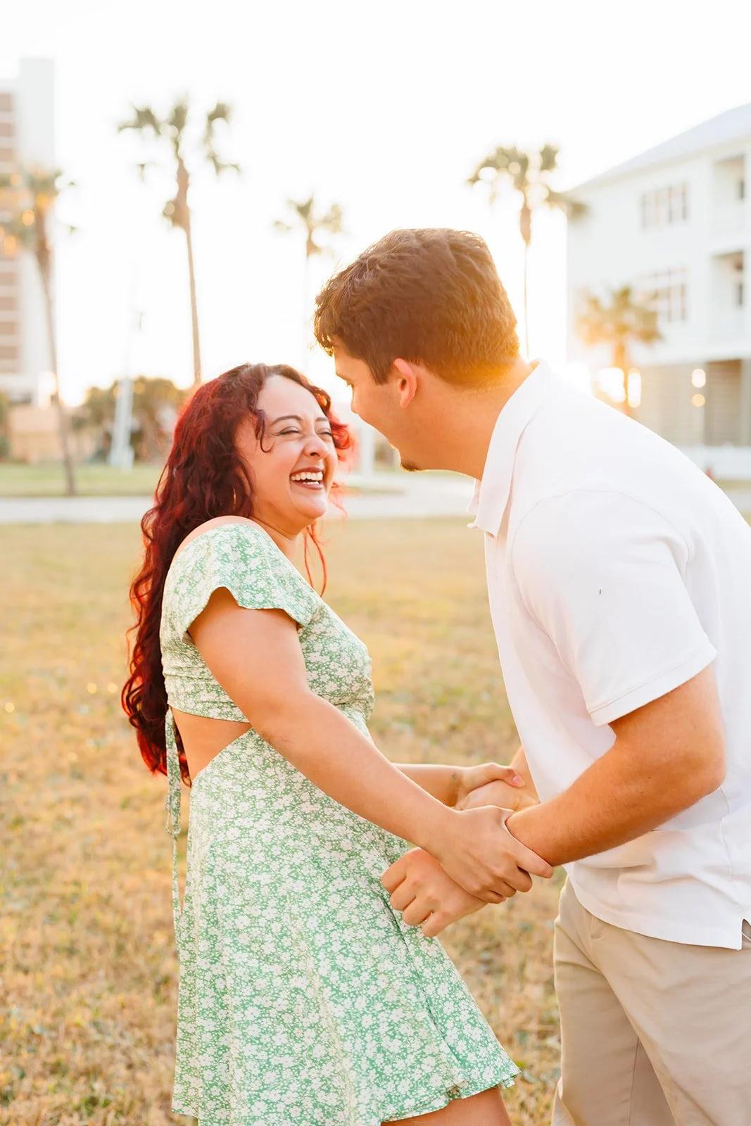 A laughing woman and a man holding hands in a grassy park at sunset with palm trees and buildings in the background.