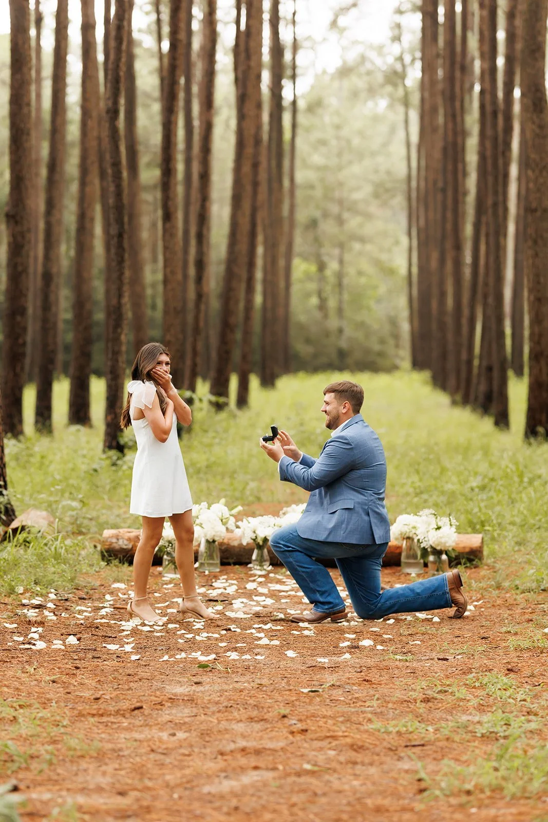 A man kneeling on one knee proposing to a woman in WG forest setting, with flower petals scattered on the ground and flowers in the background. It was captured by the best surprise proposal photographer in Houston.