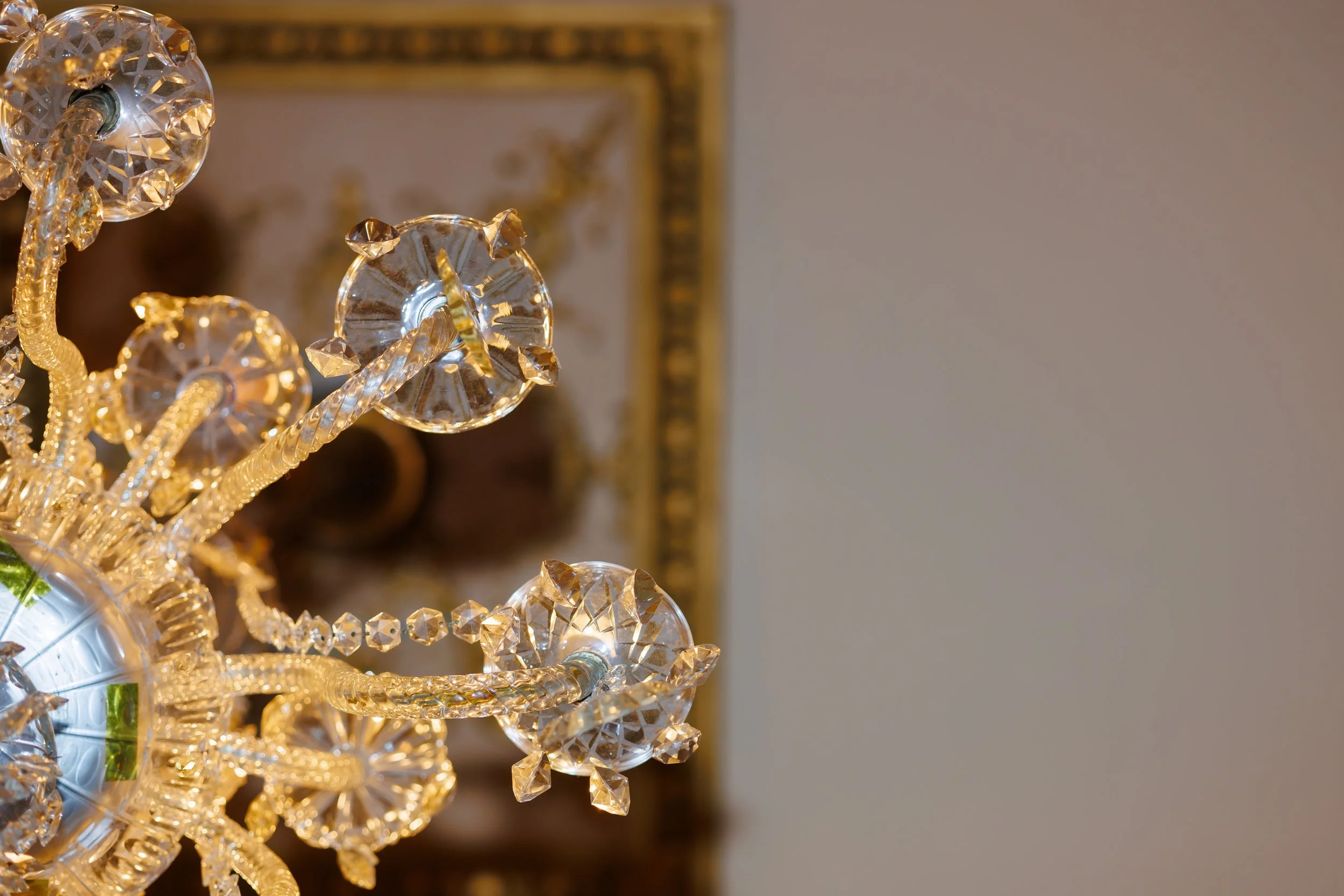 Close-up of a crystal chandelier in a luxury Houston wedding venue, with glass arms, hanging crystals, and a blurred ornate gold-framed mirror in the background.