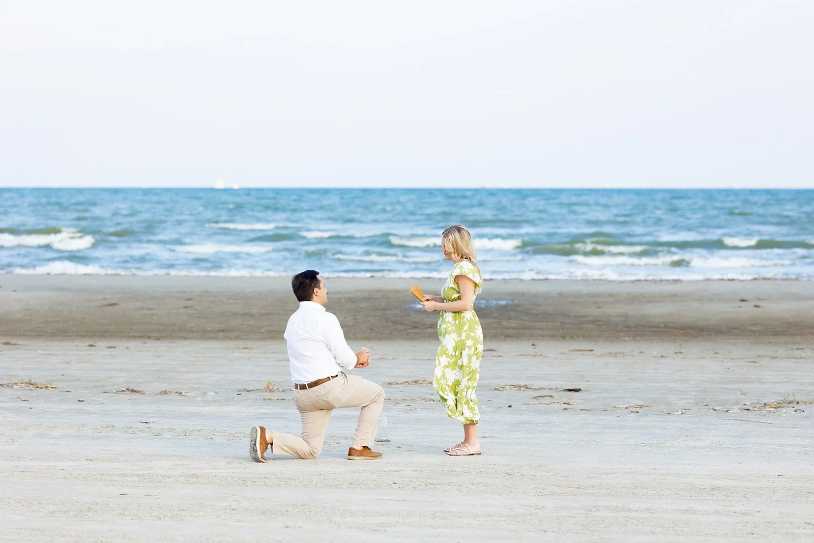 A man proposing to a woman on the beach with the ocean in the background, man kneeling with a ring box and woman standing in a yellow floral dress holding a bouquet of flowers taken by the best surprise proposal photographer in Galveston, Tx.
