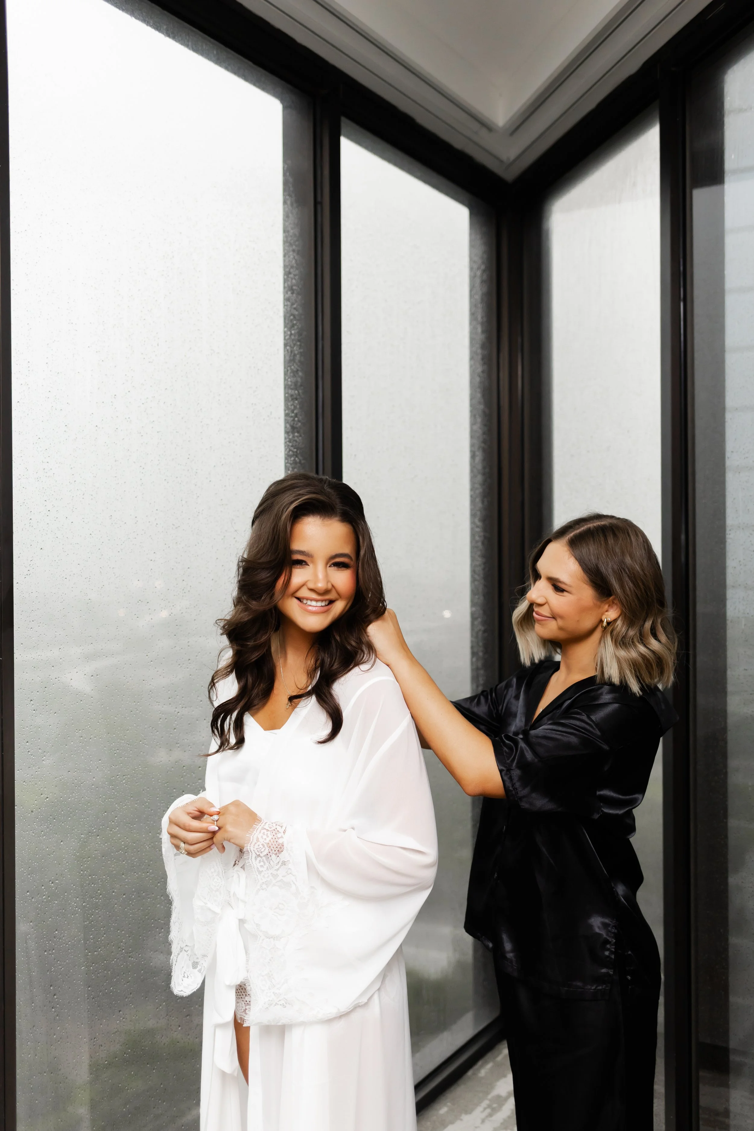 Two women, one in a white robe with lace details and the other in a black satin robe, smiling and getting ready in front of a rain-streaked window by Wild Ivy Creative Photography with Wild Ivy Creative Photography