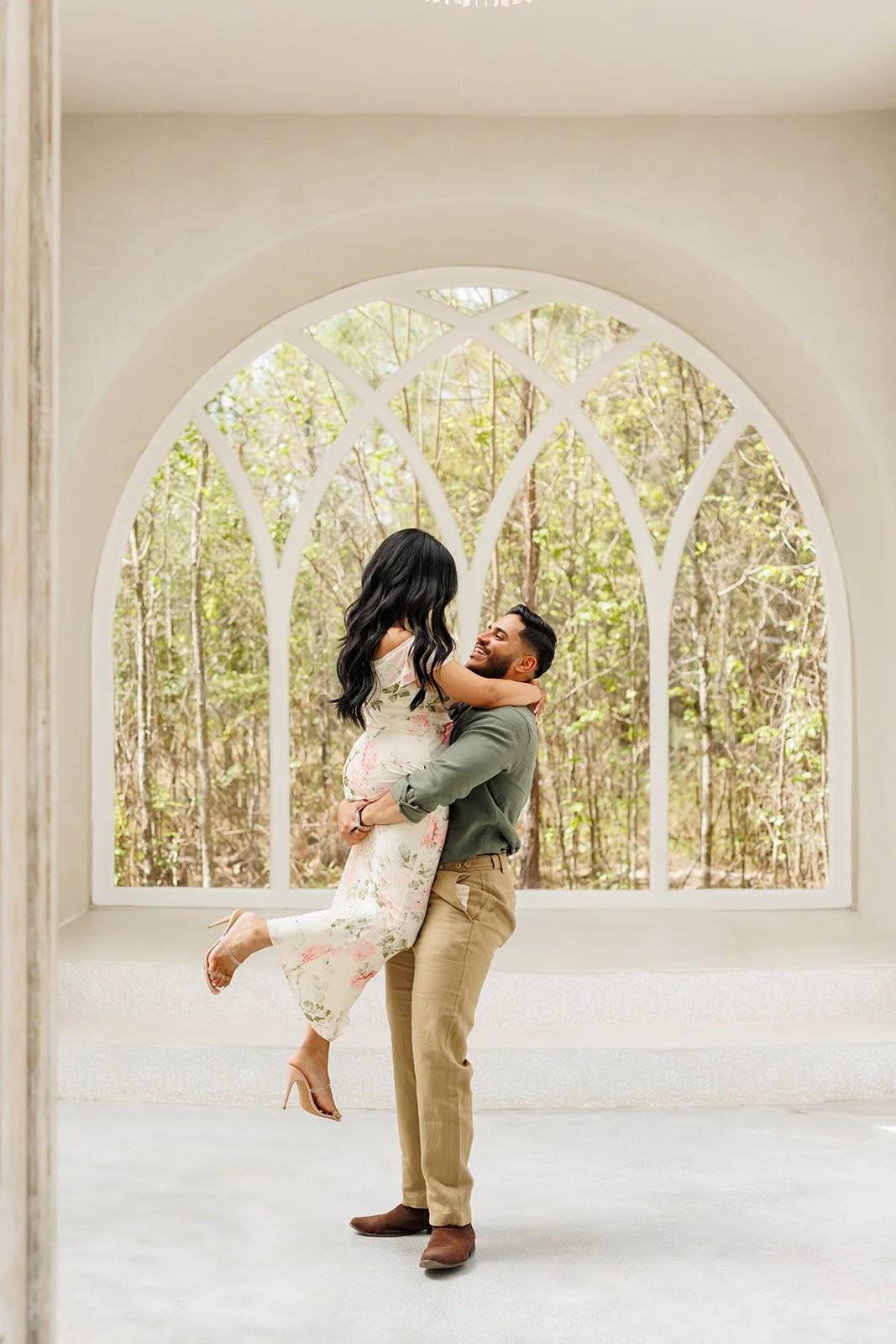 Playful indoor engagement session with a man lifting his fiancée near a large arched window in a bright space with tree views at The Rosewood in The Woodlands.