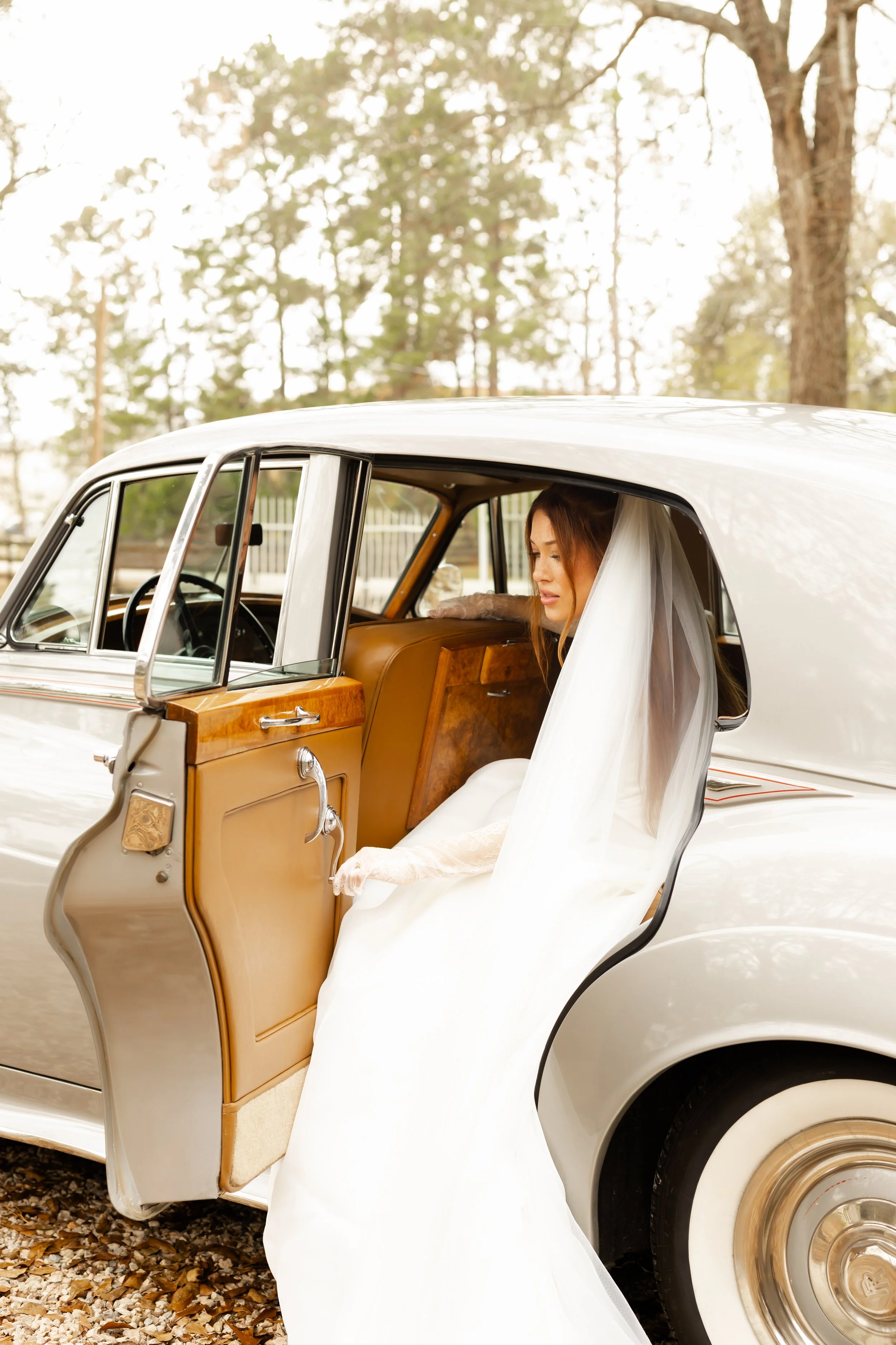 A bride in a white wedding dress and veil sitting in a vintage cream-colored car with the door open, outside with trees in the background.