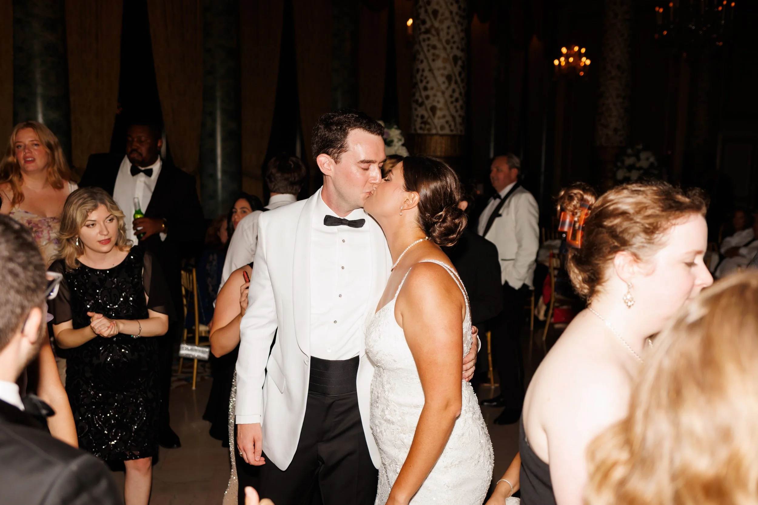 Bride and groom kissing at their Houston wedding reception, surrounded by family and friends in elegant evening attire, with warm golden light and celebratory atmosphere.