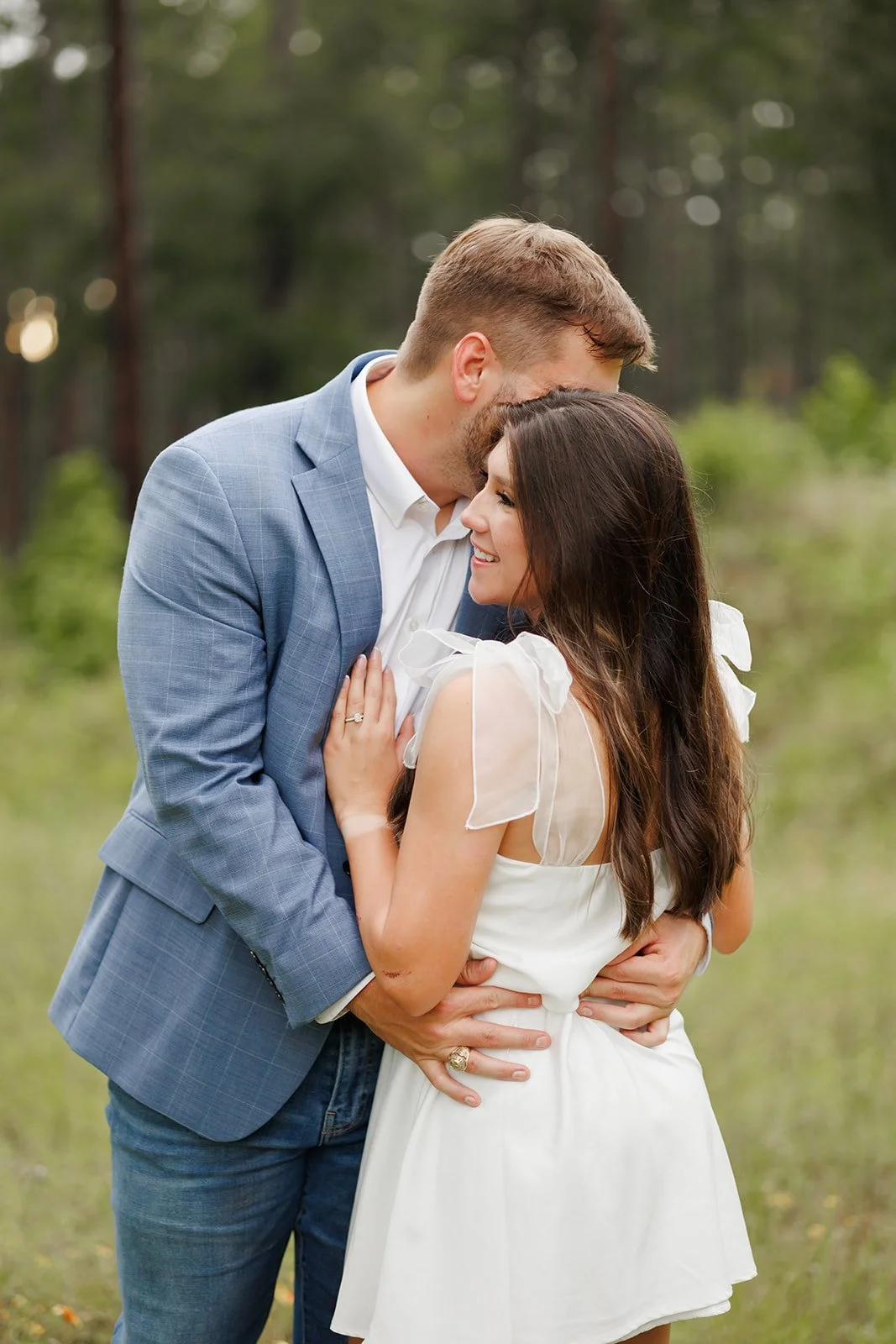 A happy couple embracing outdoors in a forest, with the man in a light blue suit and the woman in a white dress with bow-tied shoulders, smiling at each other.