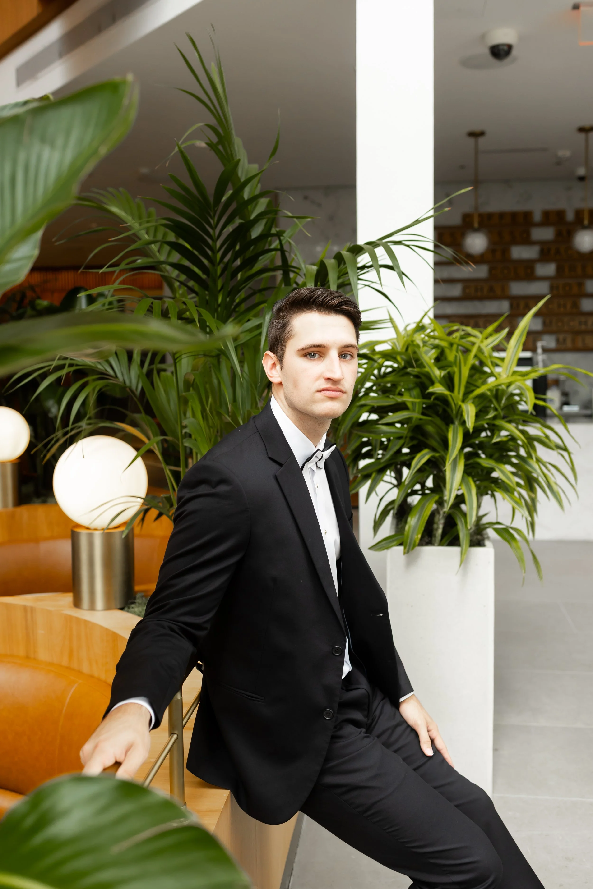 A young man dressed in a formal black tuxedo with a bow tie sitting on a wooden bench in a modern indoor setting with large green potted plants around him photographed by Wild Ivy Creative Photography