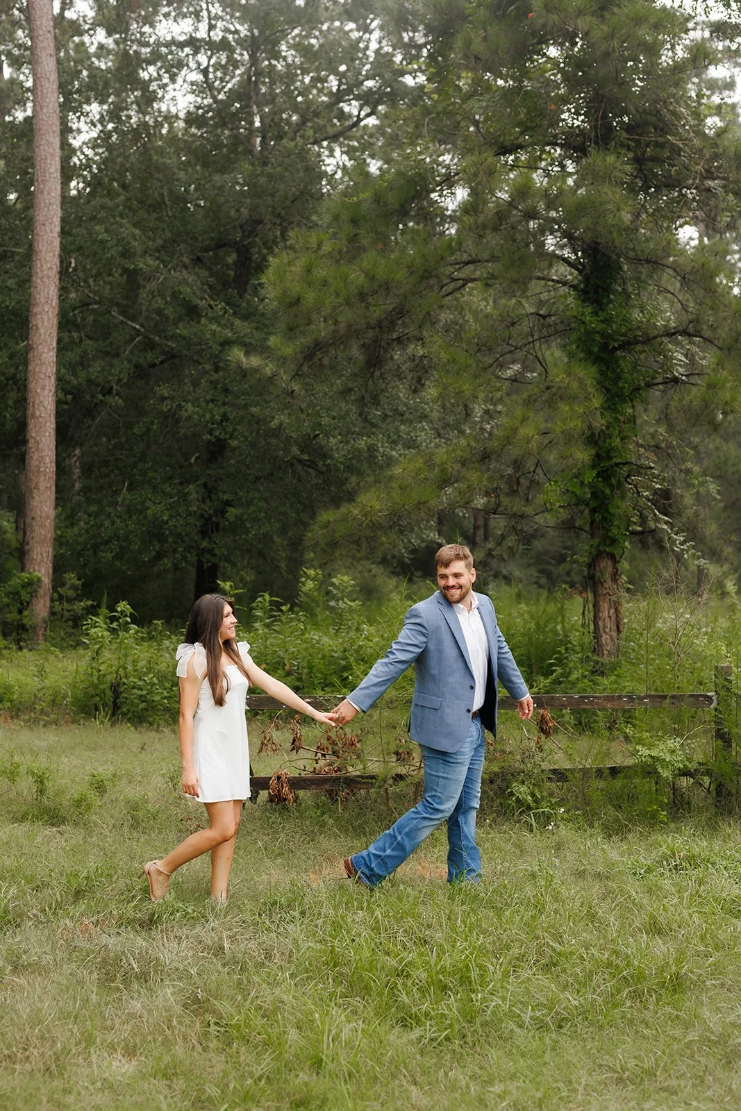 Outdoor engagement session in Houston at WG Jones State Parkwith a couple smiling as they walk hand-in-hand through a grassy, tree-lined field — photographed by Wild Ivy Creative.