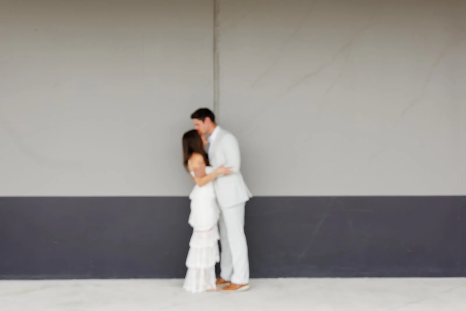 A couple in white clothing closely embracing against a two-tone gray wall.