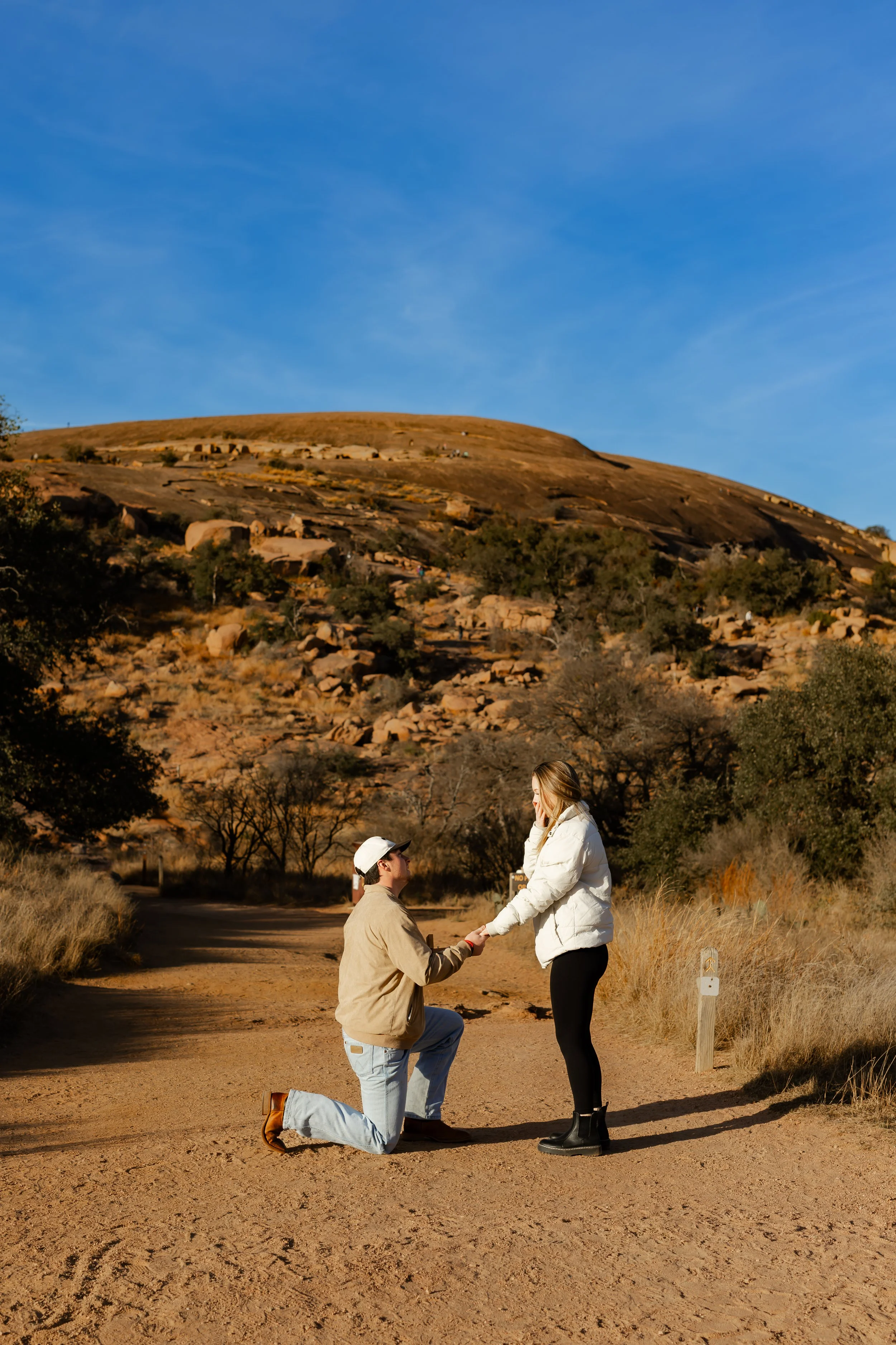 A man proposal a woman on a dirt path in a hilly, rocky landscape during daylight. The man is kneeling, holding the woman's hand, as she looks surprised. Both are dressed casually, with the woman wearing a white jacket and black pants, and the man in