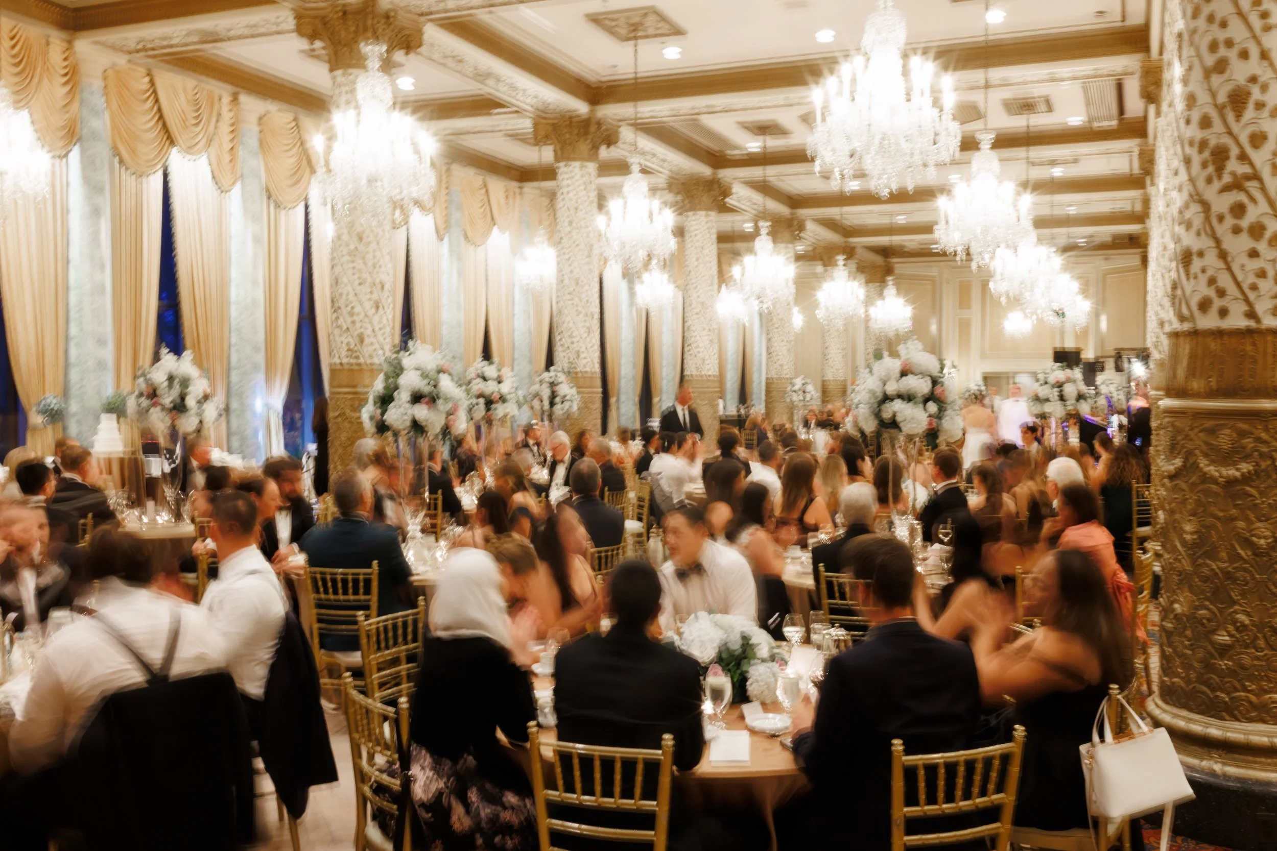Grand banquet hall in The Woodlands, Texas wedding venue, featuring gold Chiavari chairs, lavish floral arrangements, crystal chandeliers, and draped floor-to-ceiling curtains filled with guests in formal wear.