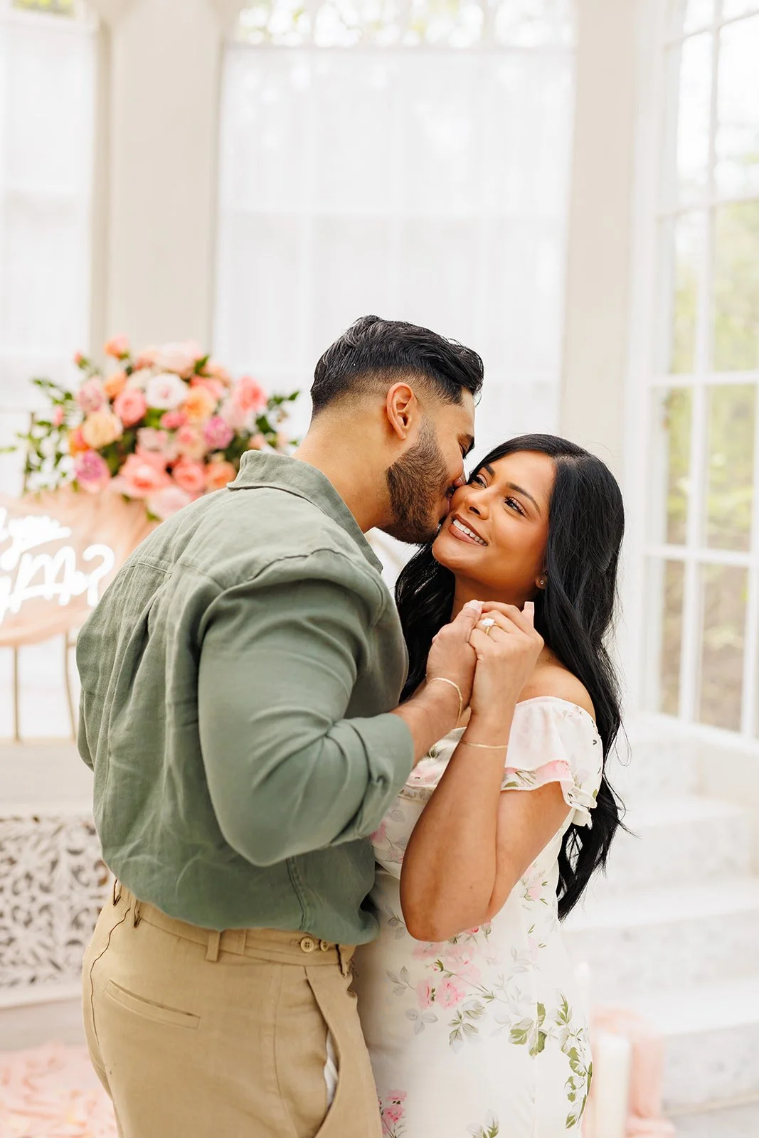 A couple shares a romantic moment during a proposal celebration indoors, with a bouquet of pink flowers in the background.