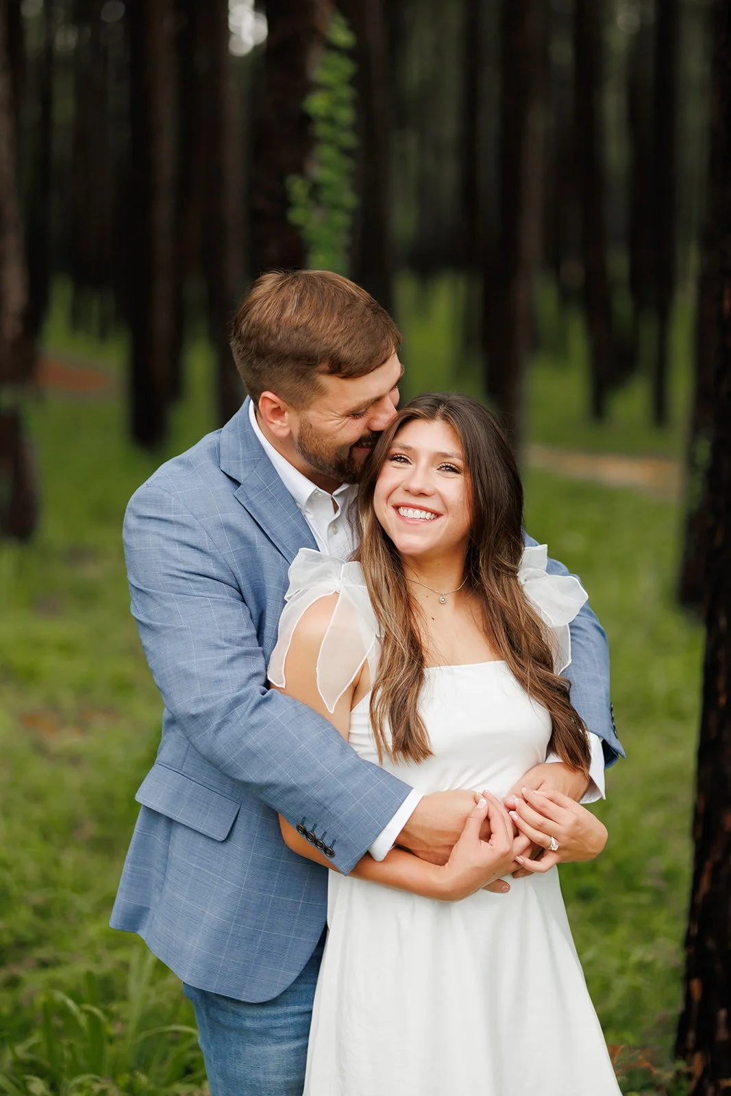 Elegant forest engagement session featuring a couple embracing, with the man in a blue suit and the woman in a white gown.