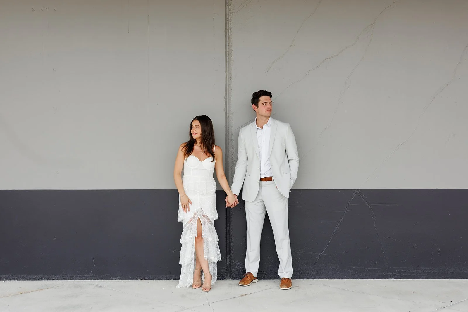 A woman and man in white formal attire holding hands while leaning against a two-tone gray wall.