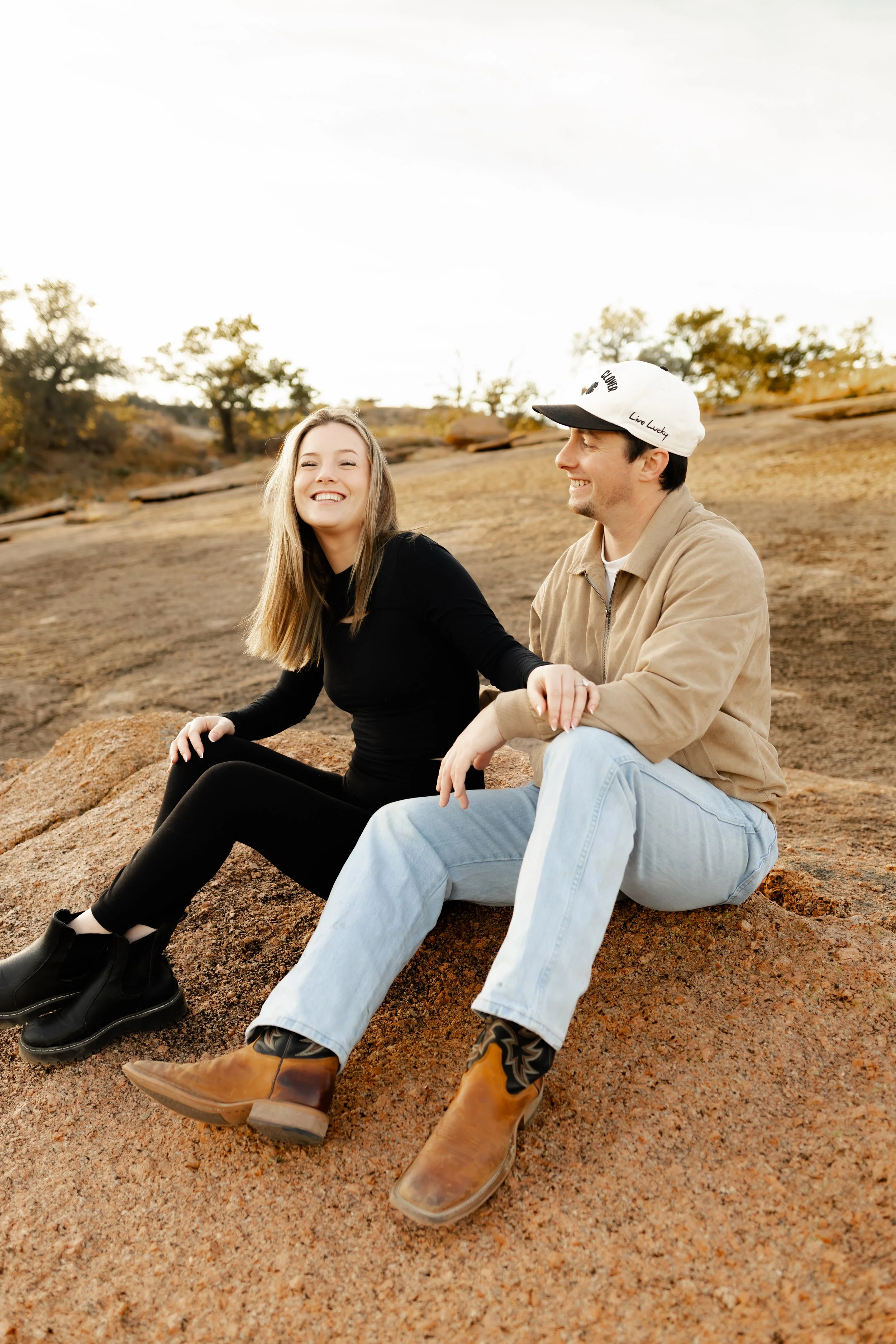 A young woman and man sitting on a rock outdoors, smiling and holding hands, during daytime with a natural landscape background Houston Engagement Proposal Photographer.