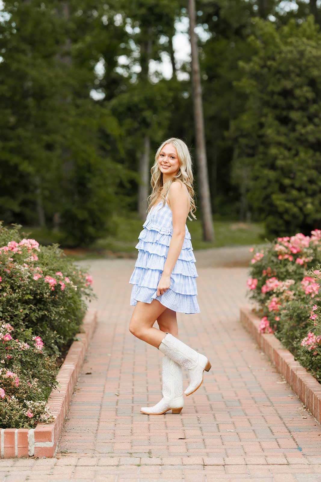 A young woman with long blonde hair wearing a blue and white ruffled dress and white cowboy boots stands on a brick pathway surrounded by pink roses and green trees, smiling at the camera at Mercer Botanic Gardens for senior portraits