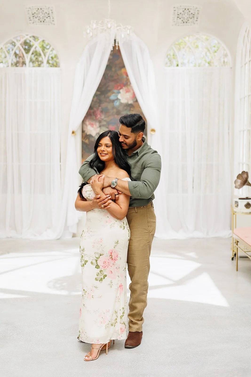 A couple sharing a warm embrace in a decorated indoor space, with white draped curtains and floral artwork in the background during their Houston Surprise Proposal Photography Session.