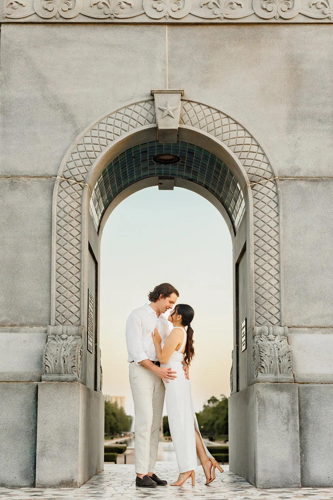 A couple dressed in white standing close together beneath an ornate stone archway, outdoors, at sunset.