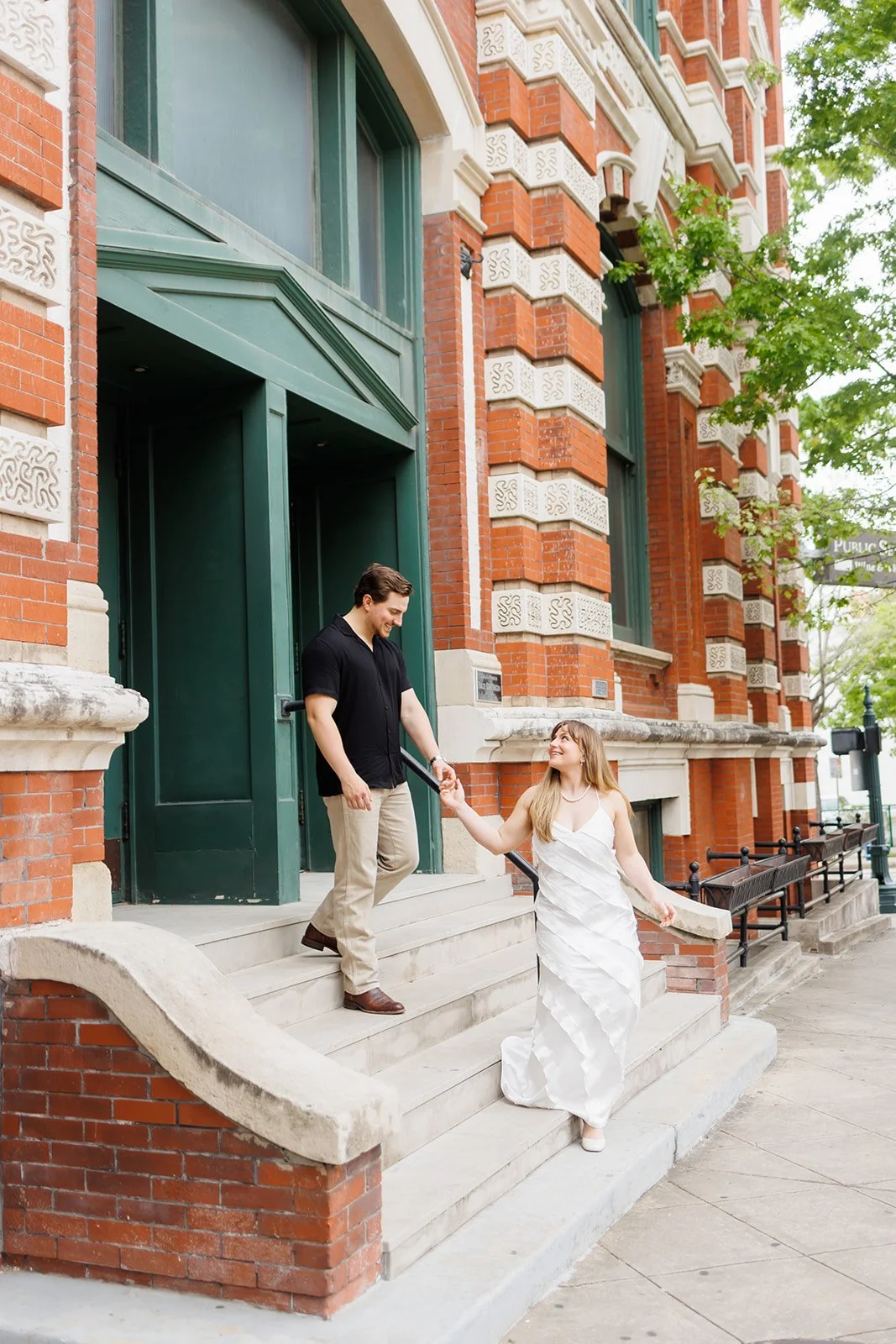 Urban engagement photo on a city street with a couple smiling and holding hands on the steps of a brick Downtown Houston building — photographed by Wild Ivy Creative.