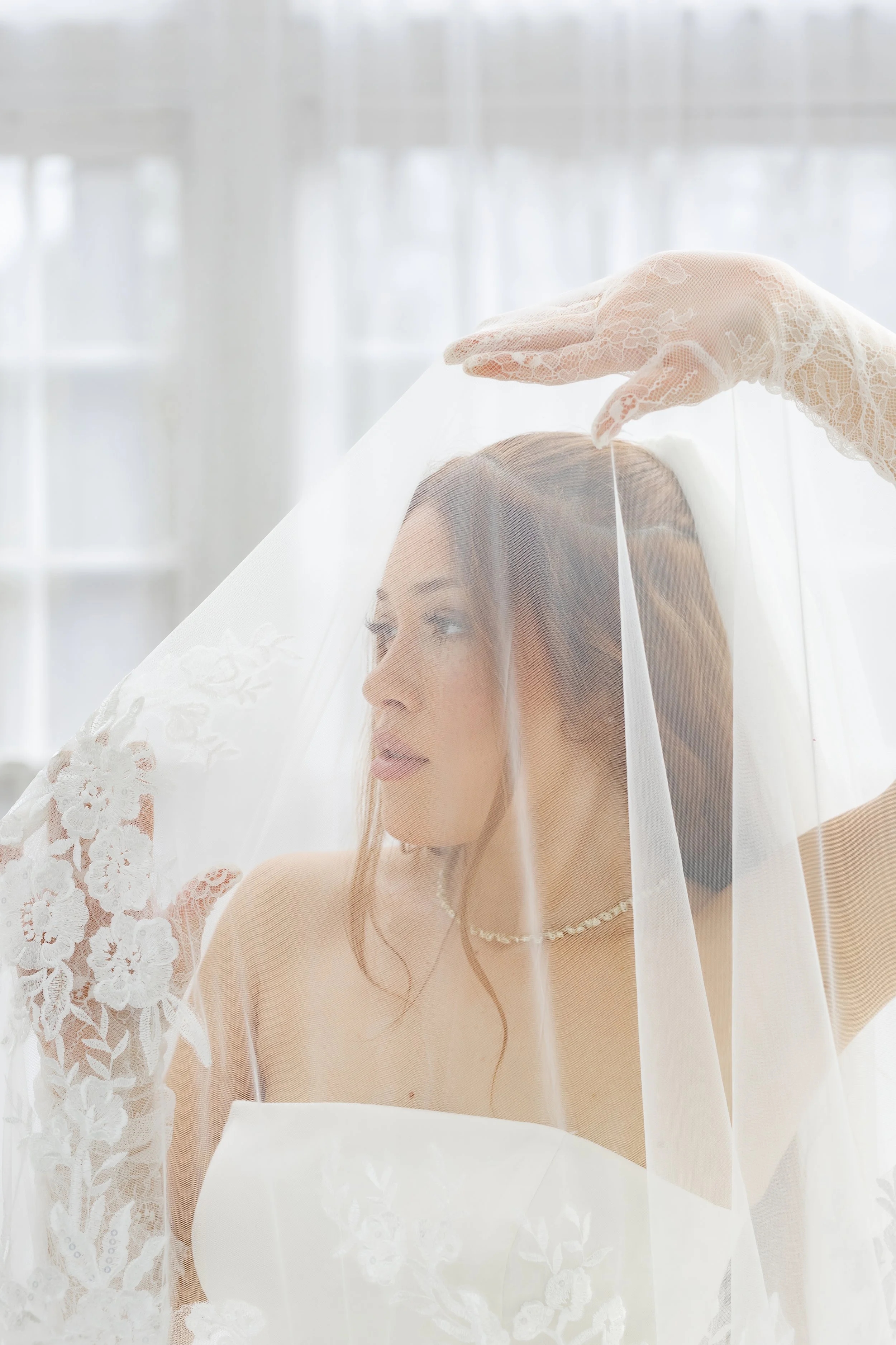 A bride with auburn hair wearing a white strapless wedding dress, lace gloves, and a pearl necklace, holding a sheer veil over her face and looking to the side indoors with a bright, soft-lit background.
