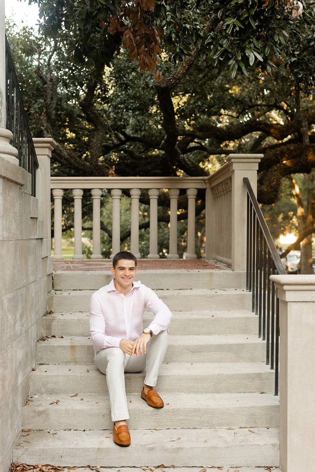 A young man sitting on steps outside, smiling, wearing beige pants, and brown shoes. Behind him are trees and a white railing, with sunlight filtering through the leaves for classic senior portraits by Wild Ivy Creative Photography at Rice University