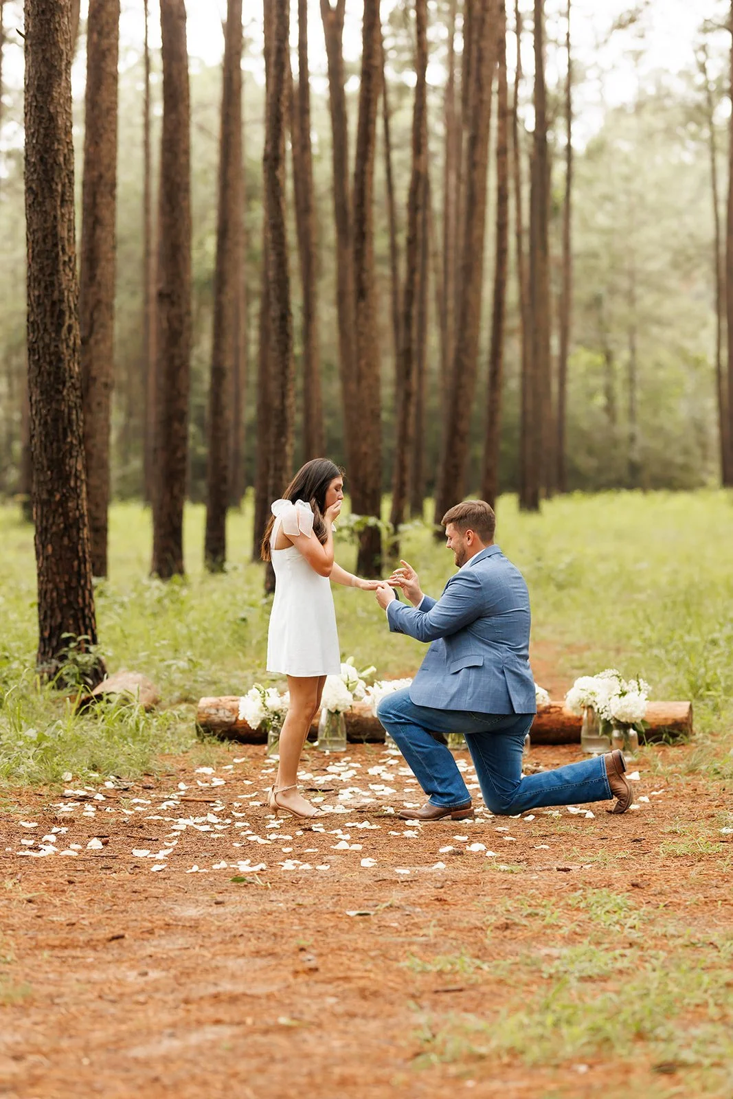 A man proposing marriage to a woman in a forest setting with flower decorations and flower petals on the ground.