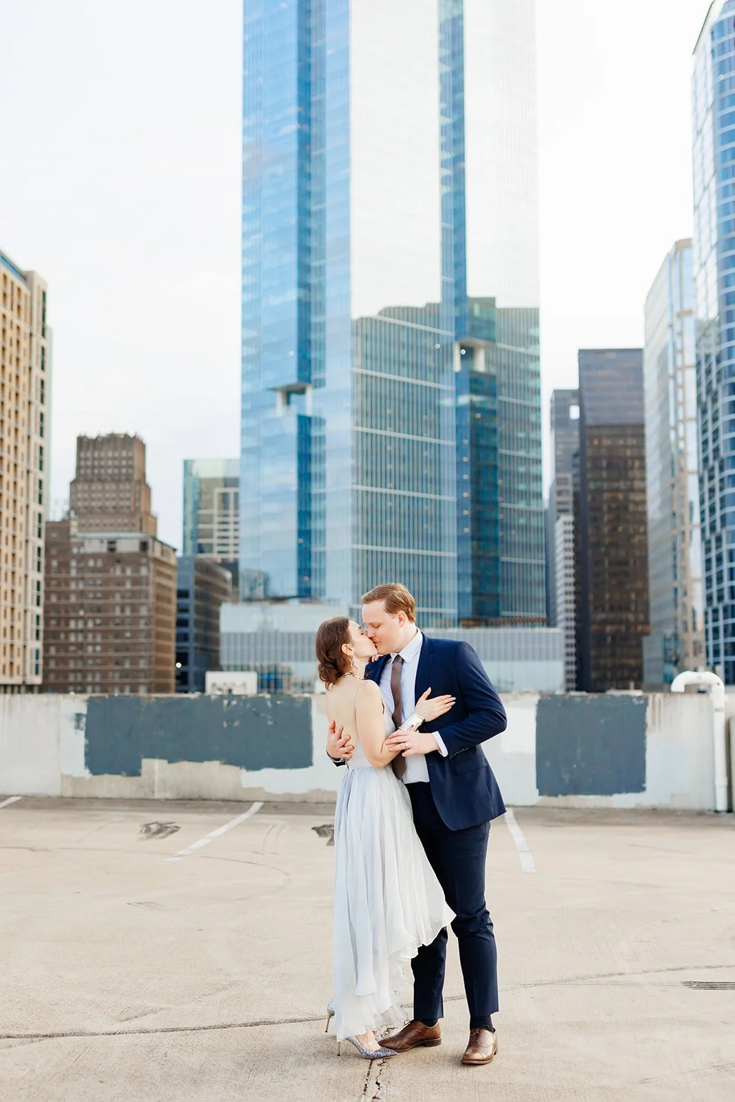 Modern rooftop engagement session in downtown Houston with a couple in formalwear sharing a kiss with the city skyline behind them — photographed by Wild Ivy Creative.