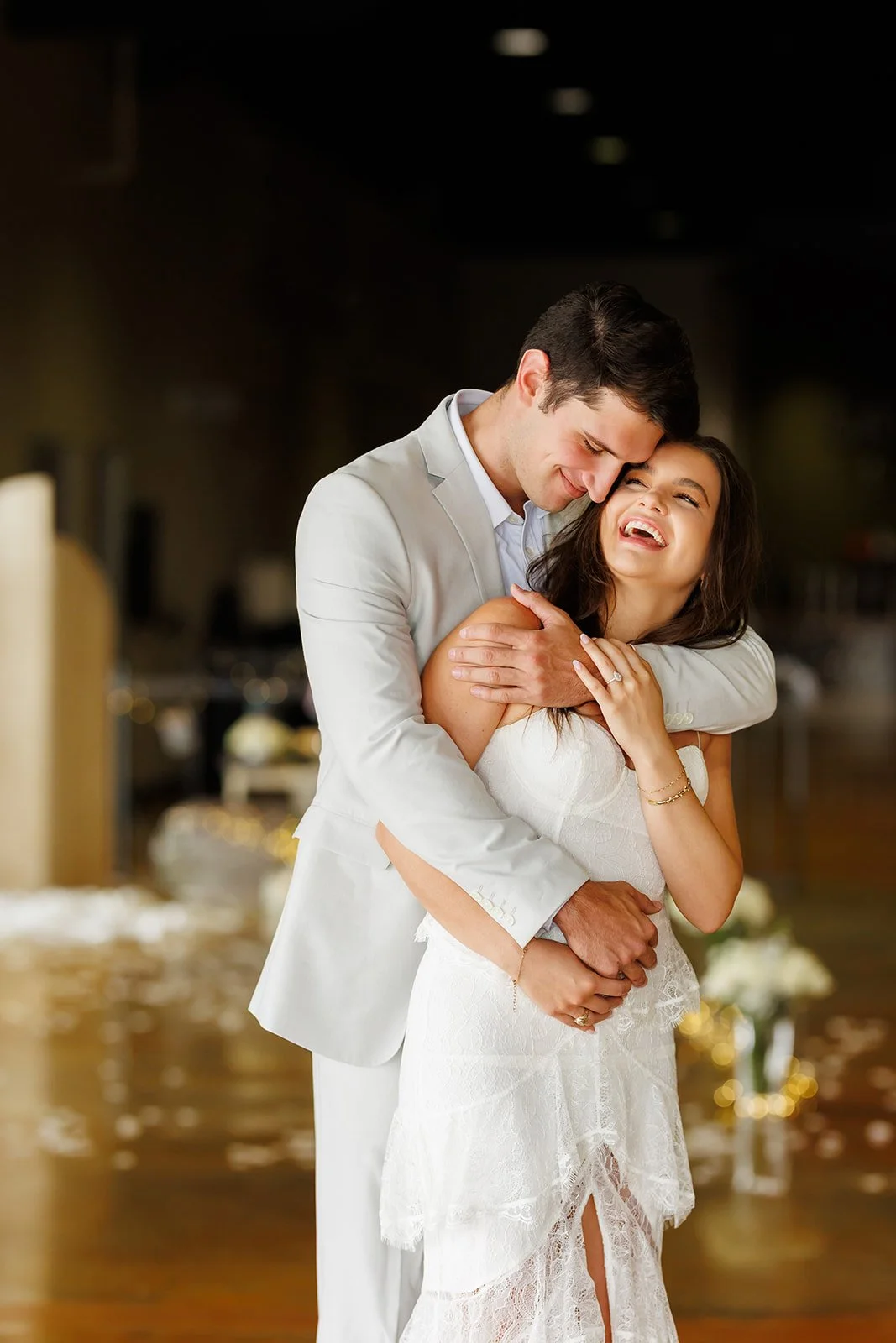 A happy couple, dressed in wedding attire, sharing an embrace and smiling at each other indoors.