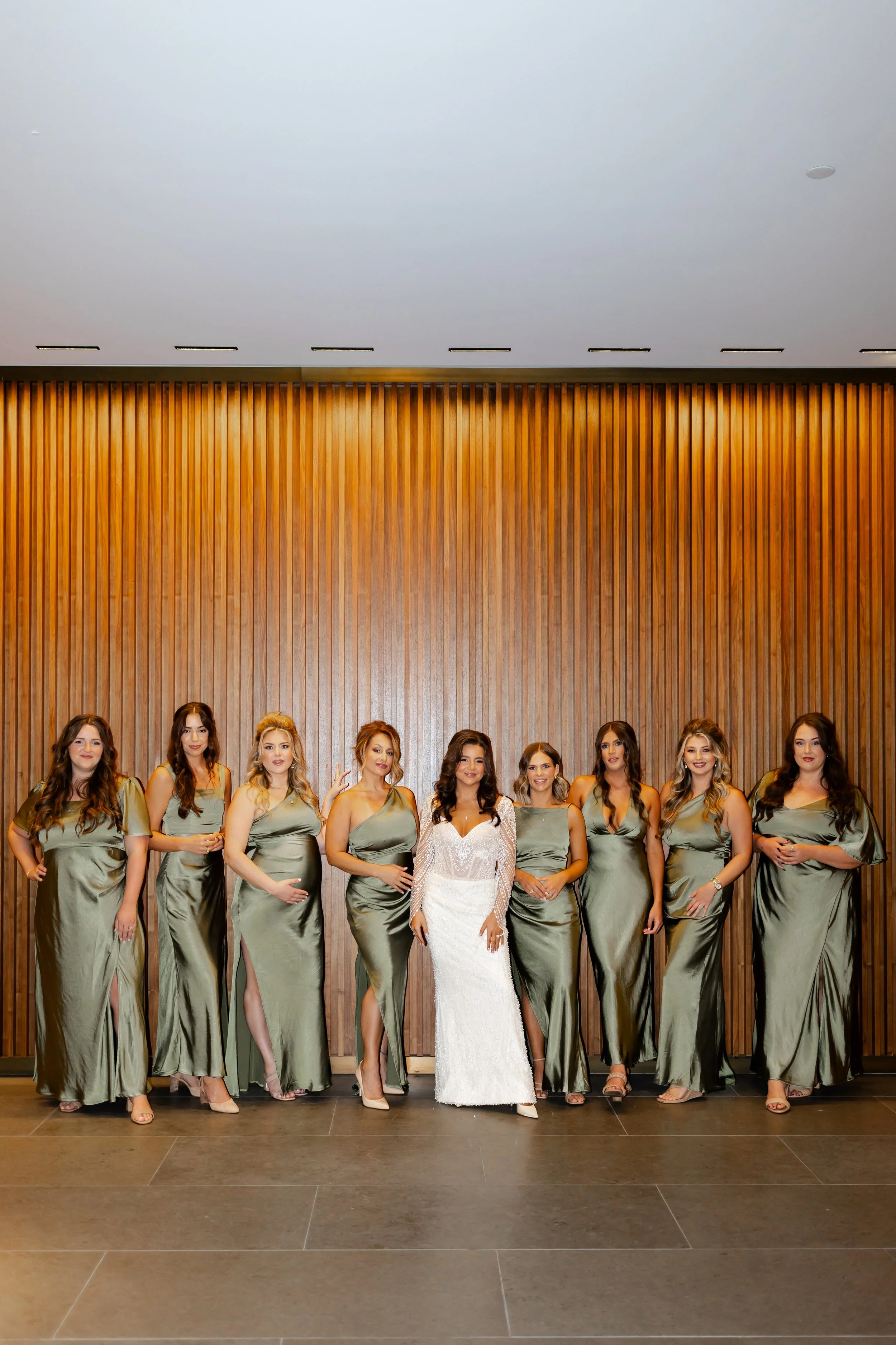 A bride and eight bridesmaids in matching olive green dresses standing in front of a wood-paneled wall at a wedding at the C. Baldwin photographed by Wild Ivy Creative Photography